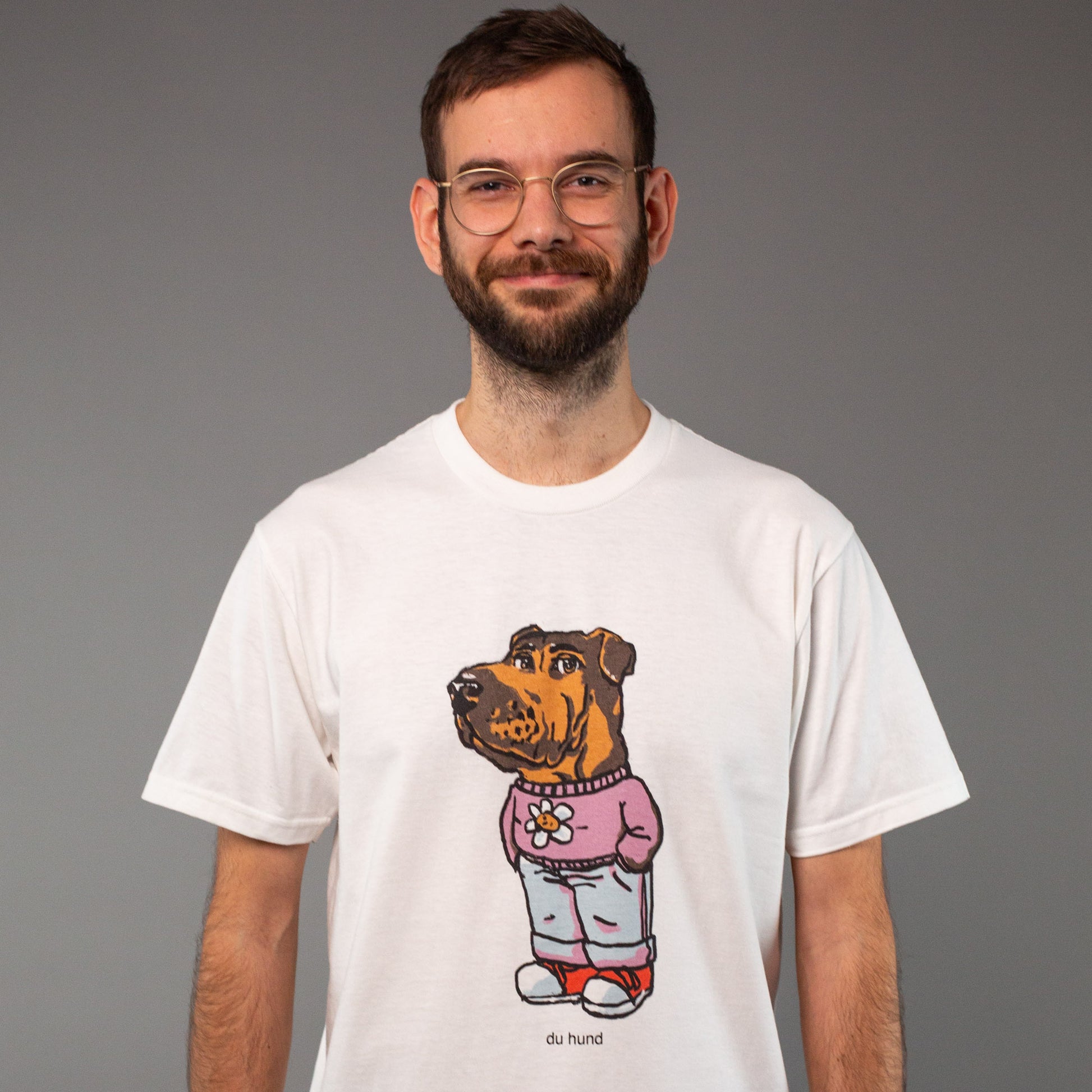 Ein Mann mit Brille und kurzem Bart trägt ein weißes T-Shirt mit einem großen, mittig platzierten Print. Das Motiv zeigt einen aufrecht stehenden Hund mit braunem Fell, der menschliche Kleidung trägt: ein pinkes Sweatshirt mit einer Blume auf der Brust, eine hellblaue Jeans und rote Sneaker. Unter dem Motiv steht der Text „du hund“. Der Mann lächelt leicht und blickt in die Kamera. Der Hintergrund ist neutral grau.