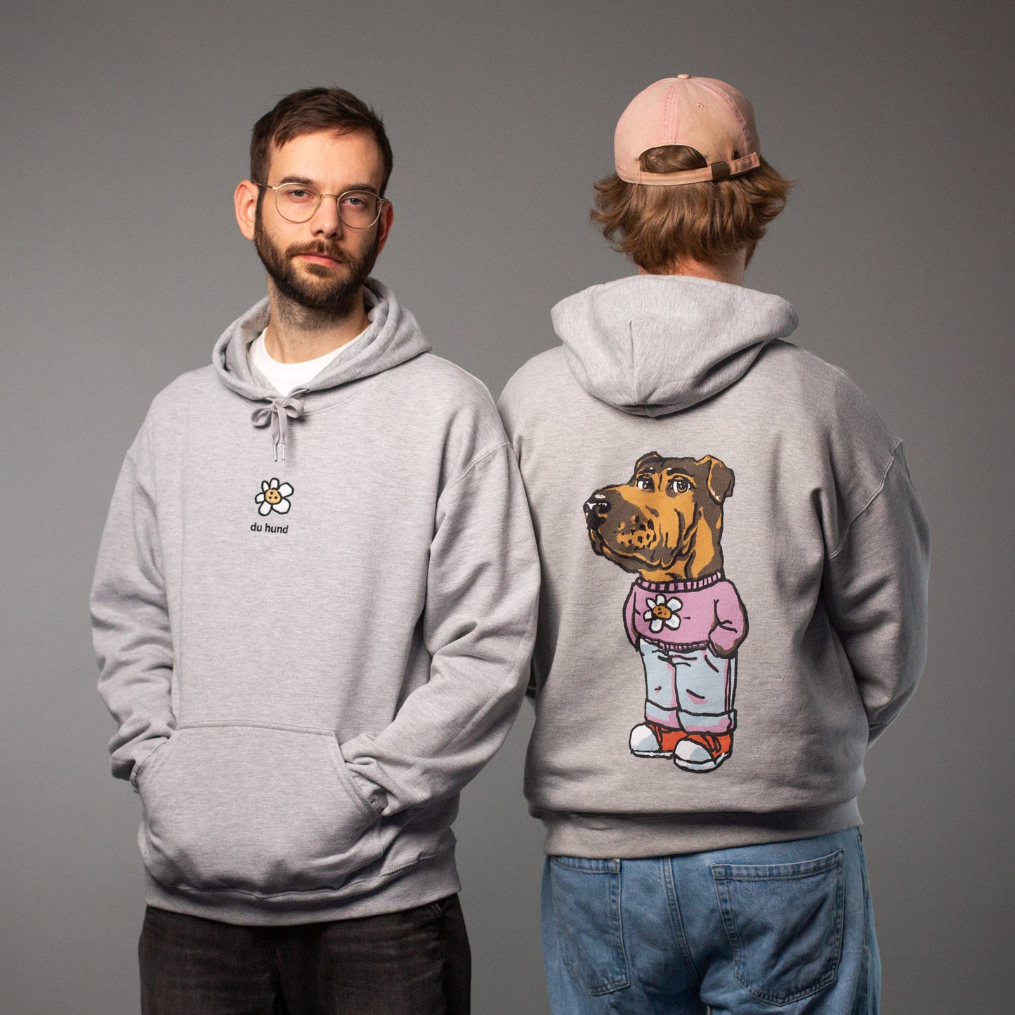 Zwei Männer tragen denselben grauen Hoodie mit „du hund“-Motiv. Der linke zeigt die Vorderseite mit einer kleinen weißen Blume mit Keks-Gesicht und dem Text „du hund“ darunter. Der rechte steht mit dem Rücken zur Kamera und zeigt den großflächigen Rückendruck: ein Hund im rosa Pulli mit derselben Blume, Jeans und Sneakern.