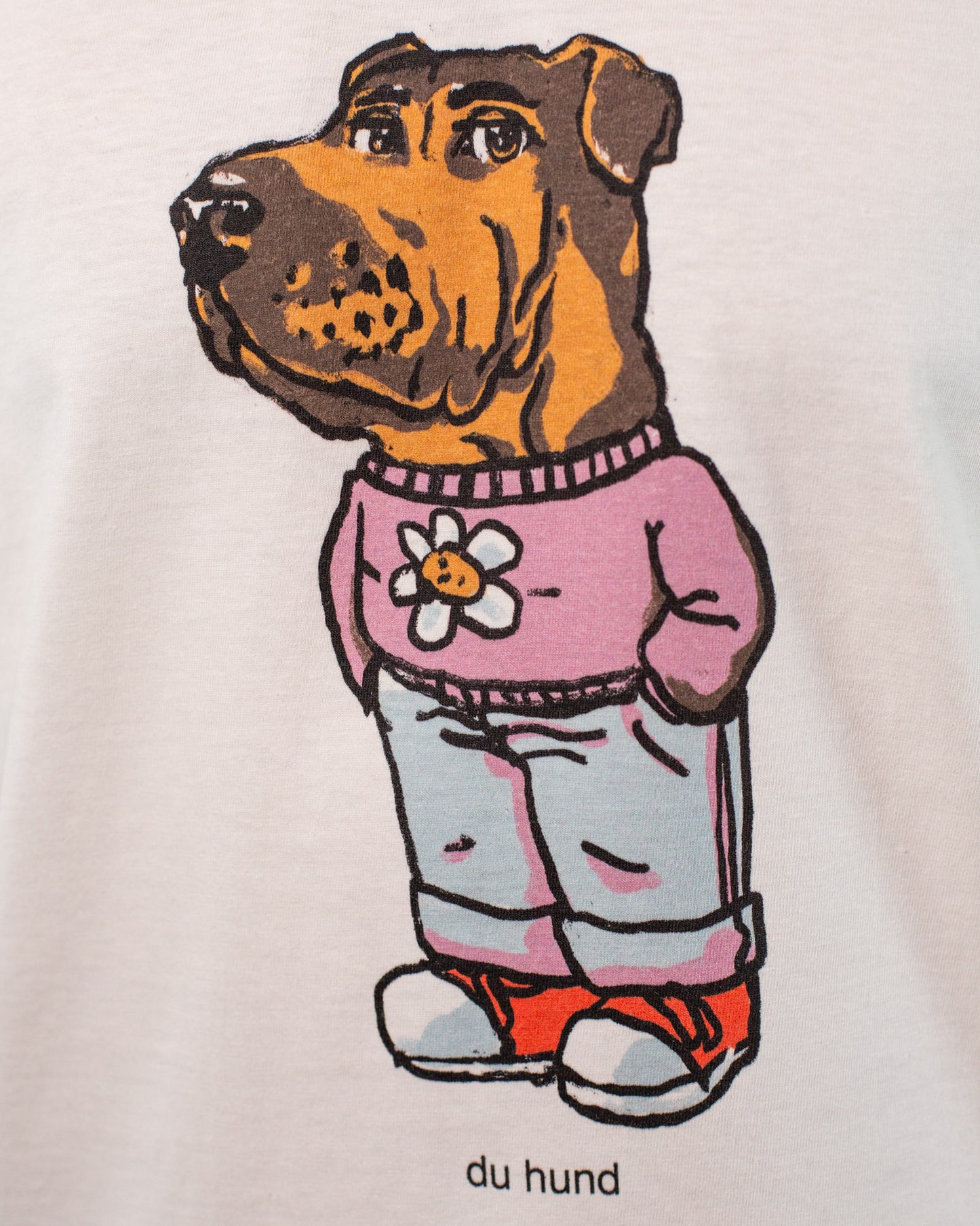 Nahaufnahme eines Drucks auf einem weißen T-Shirt. Der Print zeigt eine Illustration eines Hundes mit braunem Fell, der aufrecht steht und menschliche Kleidung trägt: ein pinkes Sweatshirt mit einer weißen Blume auf der Brust, eine hellblaue Jeans und rote Turnschuhe. Unter dem Motiv steht in schwarzer Schrift „du hund“. Die Linien des Drucks sind grob umrissen, der Stil erinnert an Comic-Grafik.