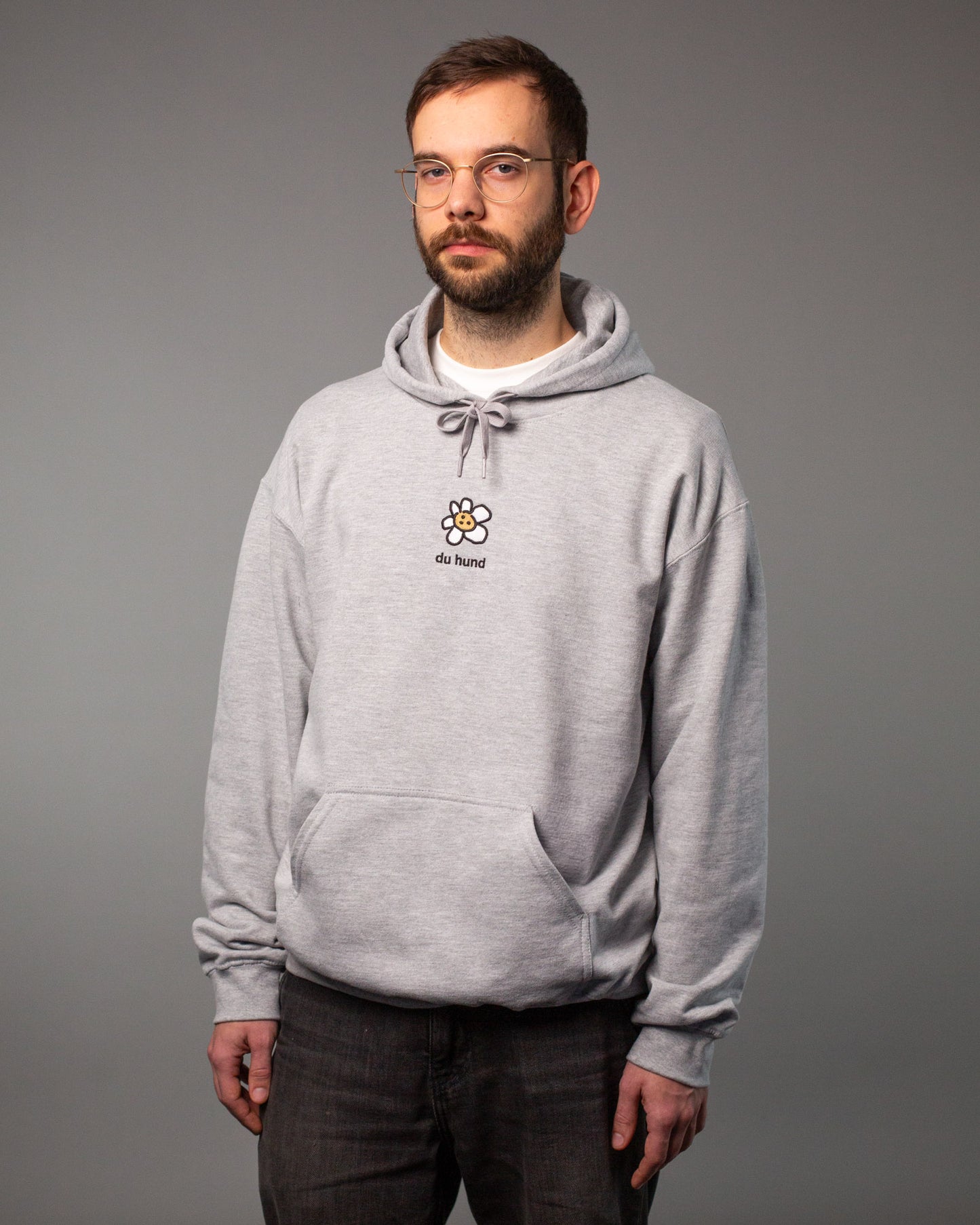 Ein Mann steht frontal zur Kamera und trägt einen grauen Hoodie. Auf der Brust befindet sich ein kleines Stickmotiv: eine weiße Blume mit einem Keks-Gesicht in der Mitte, darunter der Text „du hund“. Der Hoodie hat eine Kapuze mit Kordelzug und eine Bauchtasche.