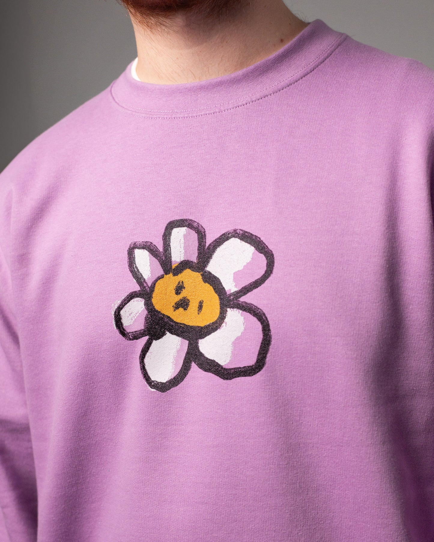 Nahaufnahme eines lilafarbenen Crewneck-Pullovers mit einem aufgedruckten Blumenmotiv. Die Blume hat weiße Blütenblätter mit schwarzer Kontur und ein gelbes Gesicht mit zwei schwarzen Punkten als Augen und einem schrägen Mund. Das Motiv sitzt mittig auf der Brust. Der Bildausschnitt zeigt den Oberkörper einer Person mit rötlichem Bart, der Kopf ist nur teilweise zu sehen.