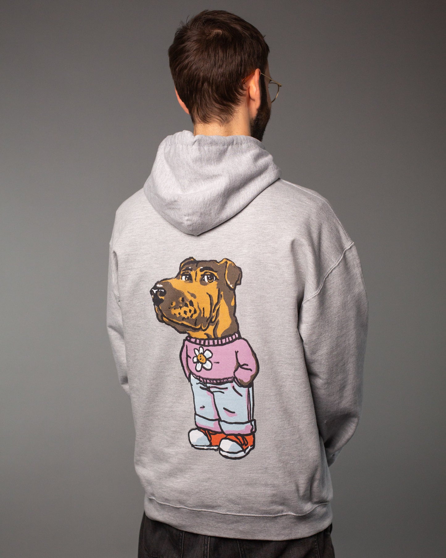 Ein Mann steht mit dem Rücken zur Kamera und trägt einen grauen Hoodie. Auf dem Rücken des Hoodies ist ein großes, buntes Motiv aufgedruckt: Ein Hund mit menschlichem Körper, bekleidet mit einem rosa Pullover mit Blümchenmotiv, hellblauer Hose und orangefarbenen Schuhen. Die Kapuze ist locker nach hinten gefallen.
