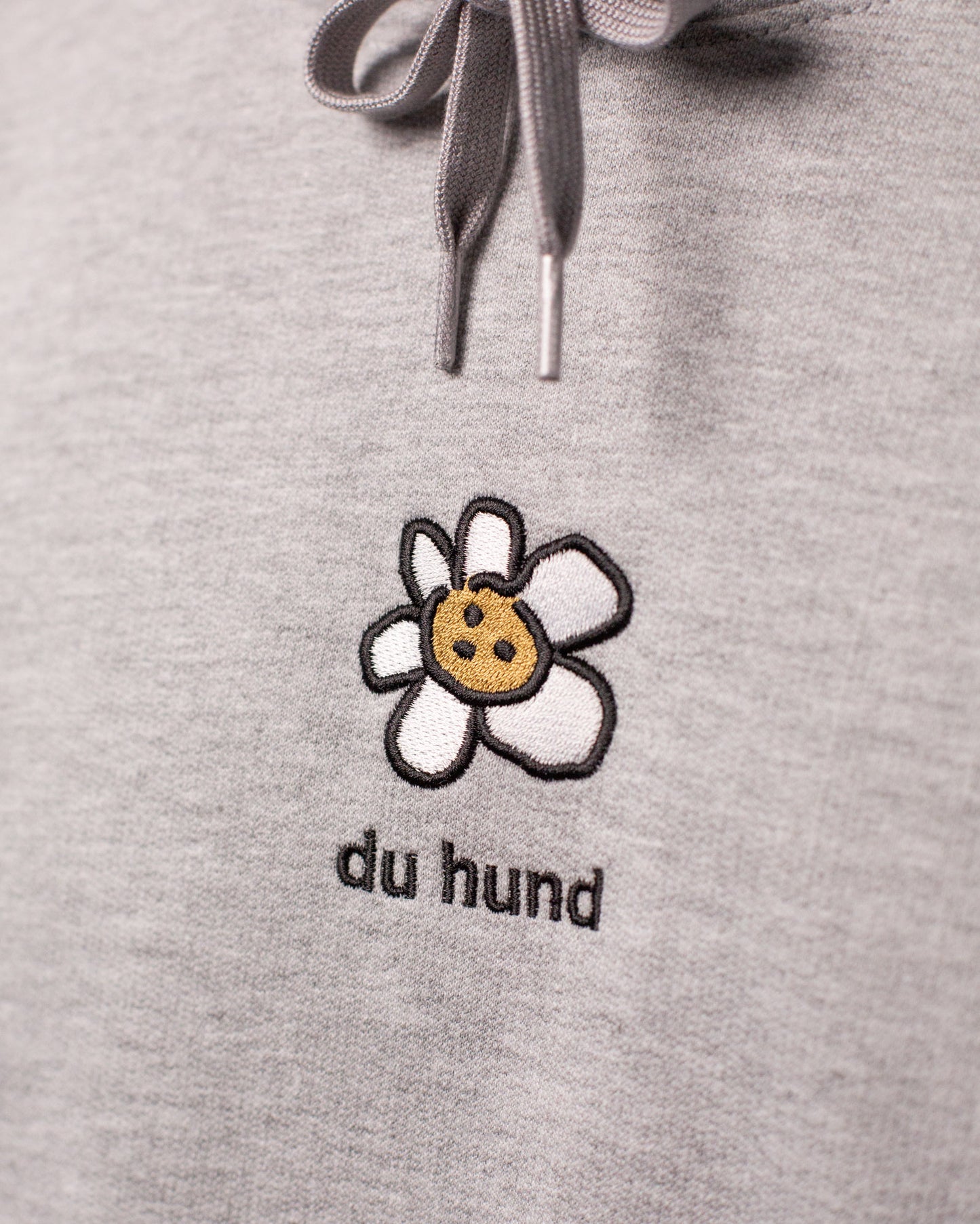 Nahaufnahme eines grauen Hoodies mit gesticktem Motiv. Zu sehen ist eine weiße Blume mit fünf Blütenblättern und einem gelben Keks mit Schokostückchen als Blütenmitte. Unter dem Motiv steht in schwarzer Schrift: „du hund“. Darüber erkennt man die Kordeln der Kapuze mit Metallenden.