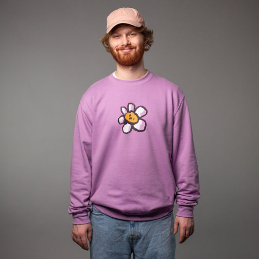 Ein junger Mann mit rotem Bart, rosa Cap und hellblauer Jeans trägt einen lavendelfarbenen Crewneck-Pullover. Auf der Brust befindet sich ein großes, zentriertes Printmotiv: eine stilisierte weiße Blume mit einem gelben Cookie-Gesicht in der Mitte. Der Hintergrund ist neutral grau, das Motiv steht klar im Fokus.