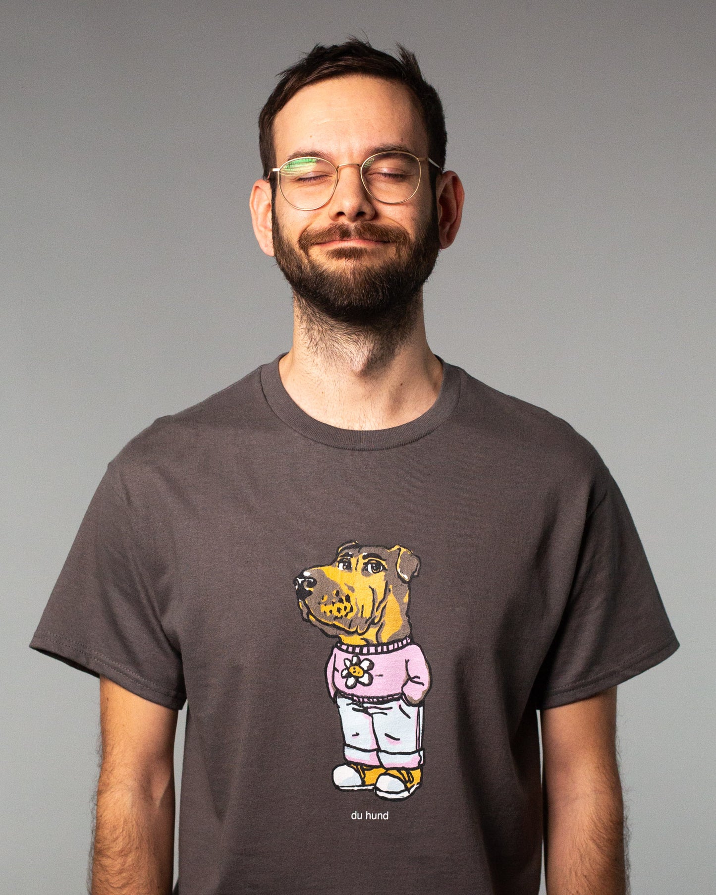 Anthrazitfarbenes T-Shirt mit zentralem Print auf der Vorderseite. Der Aufdruck zeigt eine humorvoll gezeichnete Figur mit Hundekopf und menschlichem Körper, gekleidet in eine rosa Kapuzenjacke mit Blümchen-Motiv, Blaue Jeans  und gelbe Sneakers. Unterhalb der Figur steht in kleiner weißer Schrift „du hund“. Das Shirt hat eine klassische Passform mit Rundhalsausschnitt – das Design kombiniert Comic-Ästhetik mit ironischem Statement.