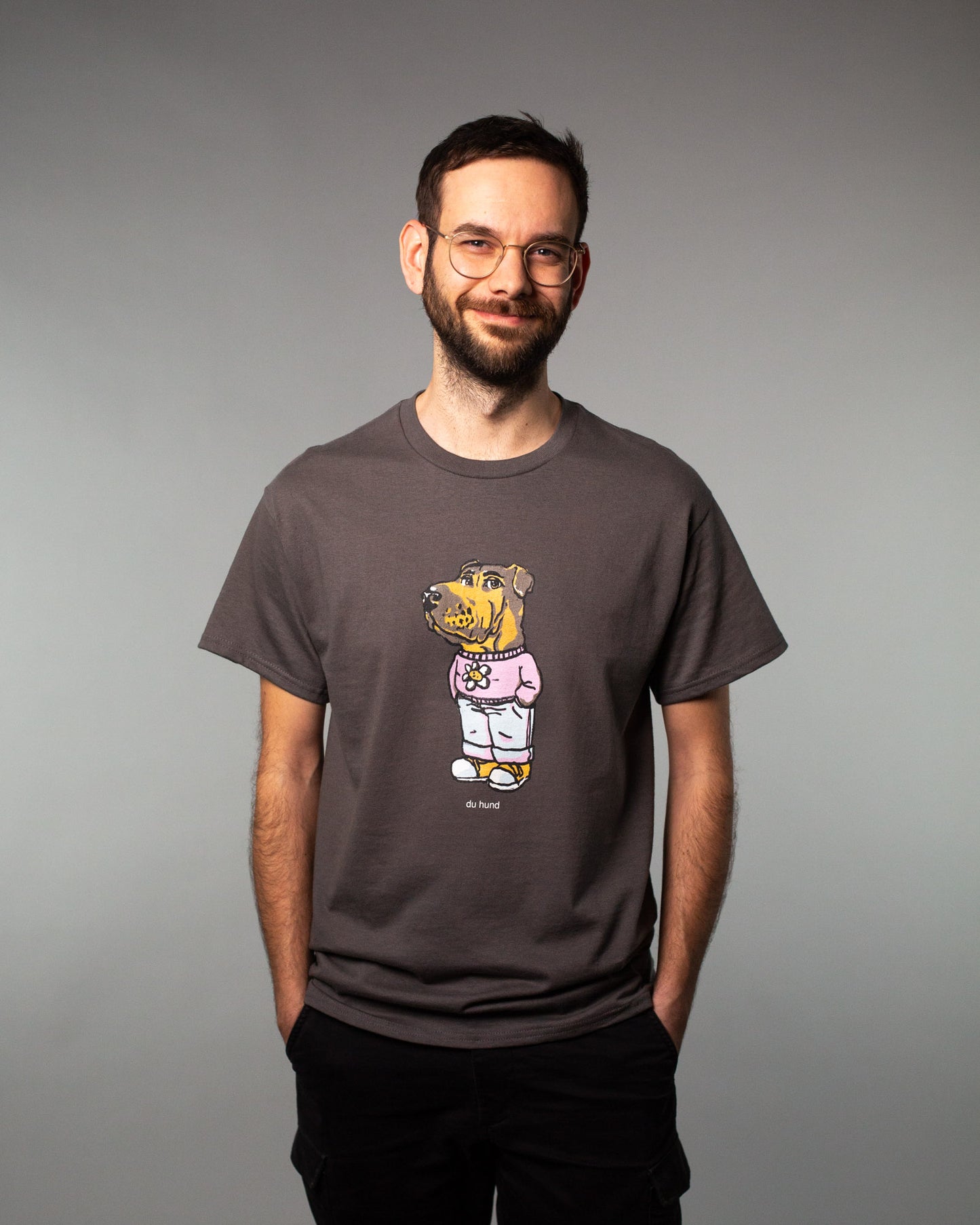 Anthrazitfarbenes T-Shirt mit zentralem Frontprint, getragen von einer Person mit leichtem Lächeln. Der Druck zeigt eine humorvolle Cartoon-Figur mit dem Kopf eines Hundes, bekleidet mit einer rosa Kapuzenjacke mit Blumenprint, Jogginghose und Sneakers. Unter dem Motiv steht in kleiner weißer Schrift der Schriftzug „du hund“. Das Shirt hat eine klassische Passform mit kurzen Ärmeln und Rundhalsausschnitt. Der Fokus liegt auf dem auffälligen, witzigen Design auf der Brust.