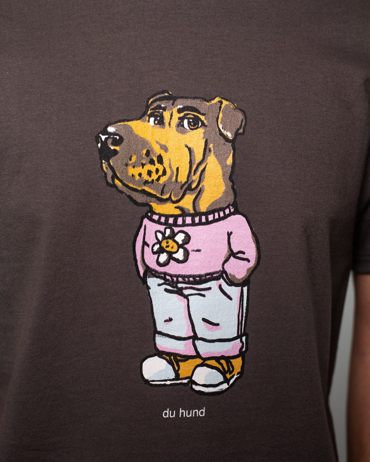 Nahaufnahme eines Aufdrucks auf einem dunkelgrauen T-Shirt. Zu sehen ist eine comichafte Figur mit dem Kopf eines Hundes und einem menschlichen Körper. Der Hund trägt eine rosa Kapuzenjacke mit einer stilisierten weißen Blume, eine hellblaue Jogginghose mit hochgekrempelten Hosenbeinen und gelbe Sneaker. Unter dem Motiv steht in kleiner weißer Schrift: „du hund“. Die Illustration ist im charmanten, handgezeichneten Stil gehalten.