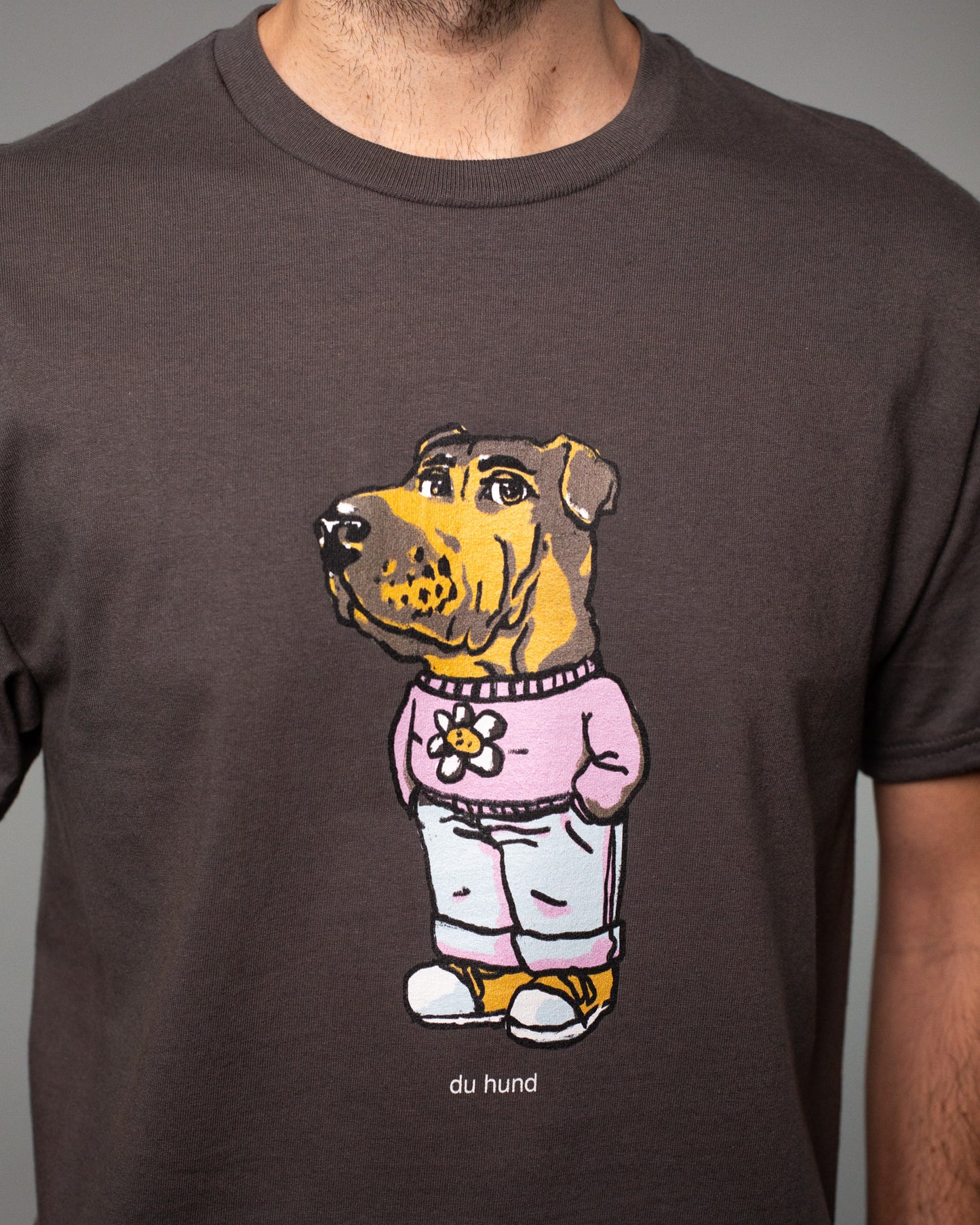 Detailaufnahme eines braunen T-Shirts mit einem großen, bunten Druck auf der Brust. Abgebildet ist eine Figur mit dem Kopf eines braunen Hundes und einem menschlichen Körper. Der Hund trägt einen rosa Pullover mit einer weißen Blume darauf, eine hellblaue Jogginghose und gelbe Turnschuhe. Unter dem Motiv steht in kleiner weißer Schrift: „du hund“. Der Zeichenstil wirkt humorvoll und illustrativ.