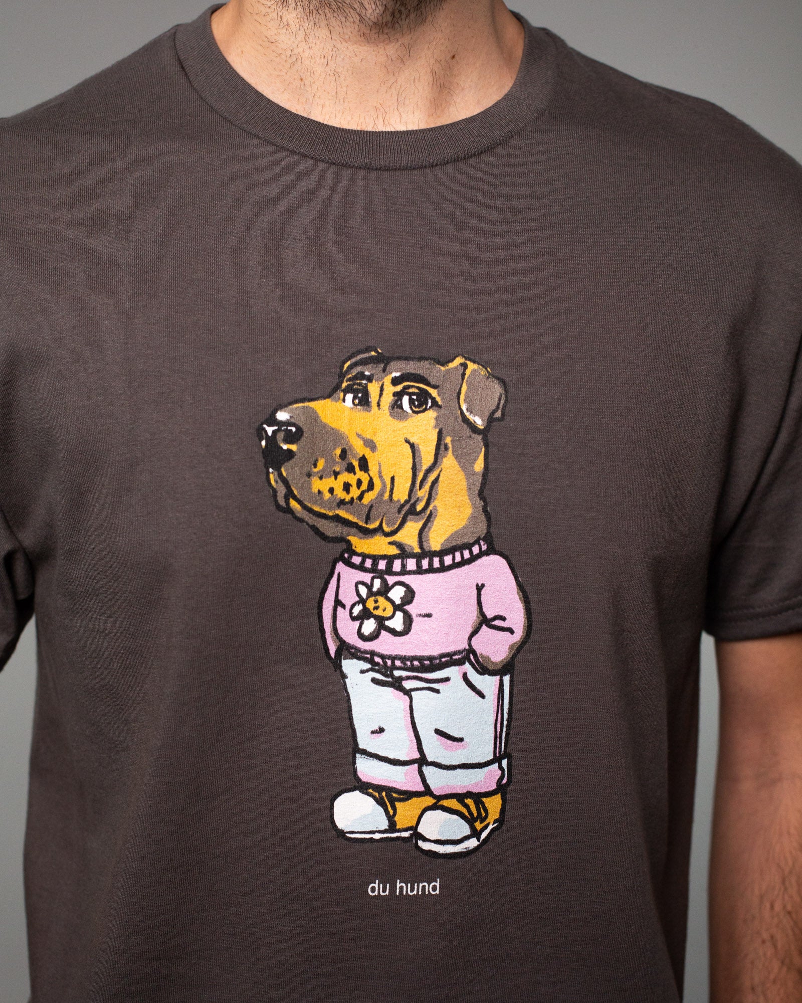 Detailaufnahme eines braunen T-Shirts mit einem großen, bunten Druck auf der Brust. Abgebildet ist eine Figur mit dem Kopf eines braunen Hundes und einem menschlichen Körper. Der Hund trägt einen rosa Pullover mit einer weißen Blume darauf, eine hellblaue Jogginghose und gelbe Turnschuhe. Unter dem Motiv steht in kleiner weißer Schrift: „du hund“. Der Zeichenstil wirkt humorvoll und illustrativ.