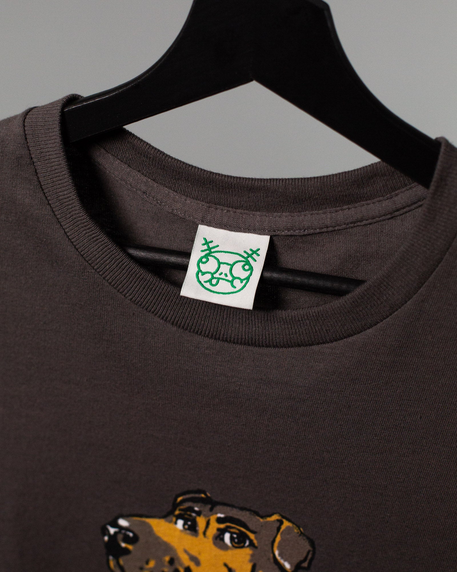 Nahaufnahme des Halsausschnitts eines braunen T-Shirts auf einem schwarzen Kleiderbügel. Am Kragen ist ein eingenähtes Etikett mit einem grünen, comicartigen Gesicht mit schielenden Augen, Brille und herausgestreckter Zunge zu sehen. Unten am Bildrand ist der obere Teil eines großflächigen Drucks mit einem gezeichneten Hundekopf zu erkennen.