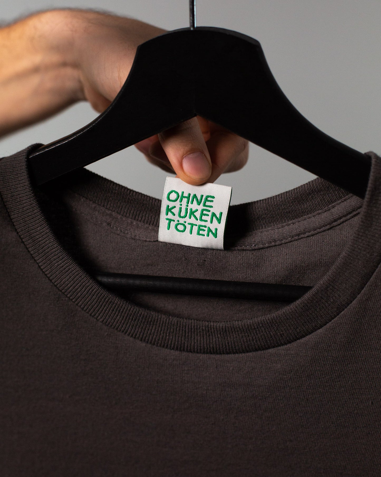 Detailaufnahme des Halsausschnitts eines braunen T-Shirts auf einem schwarzen Kleiderbügel. Eine Hand hält das eingenähte weiße Etikett nach vorn. Darauf steht in grüner Schrift: „OHNE KÜKEN TÖTEN“. Der Fokus liegt auf dem Nachhaltigkeitshinweis.