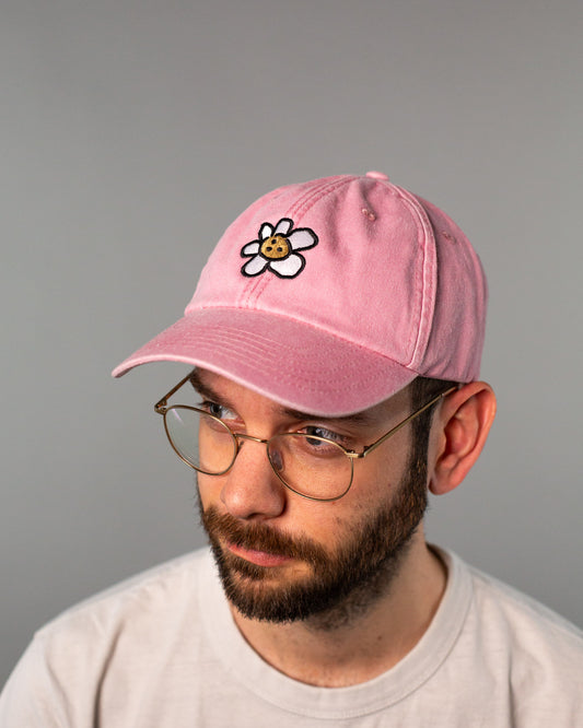 Rosa Cap aus Baumwollstoff mit gesticktem Motiv auf der Vorderseite. Das Design zeigt eine stilisierte Blume mit weißen Blütenblättern und einem Keks als Blütenmitte mit Gesicht – comicartig und verspielt. Die Cap hat eine klassische 6-Panel-Form mit gebogenem Schirm und kleinen Belüftungslöchern. Sie wird von einer Person mit Brille getragen, der Fokus liegt jedoch klar auf dem Produktdesign der Cap.
