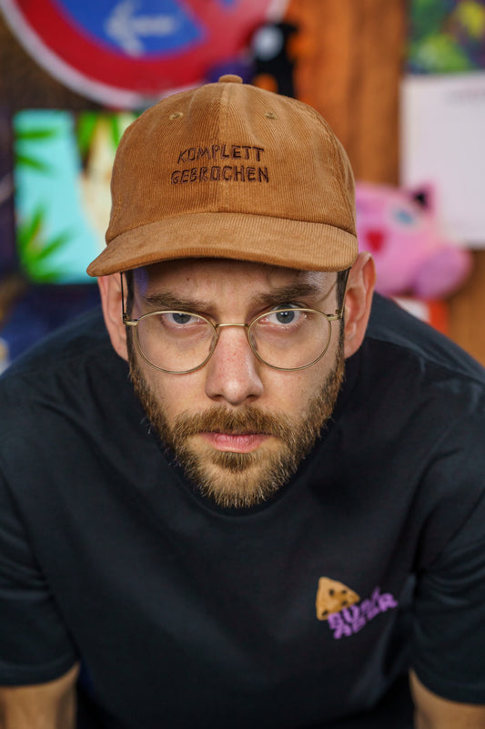 Braune Cord-Cap aus weichem Material, frontal getragen. Auf der Vorderseite ist in dunkler Schrift der Schriftzug „KOMPLETT GEBROCHEN“ sauber eingestickt. Die Cap hat eine klassische, leicht gebogene Form mit sechs Panelen und Belüftungsösen. Der Stoff wirkt strukturiert und hochwertig – ideal für einen lässigen, individuellen Look.