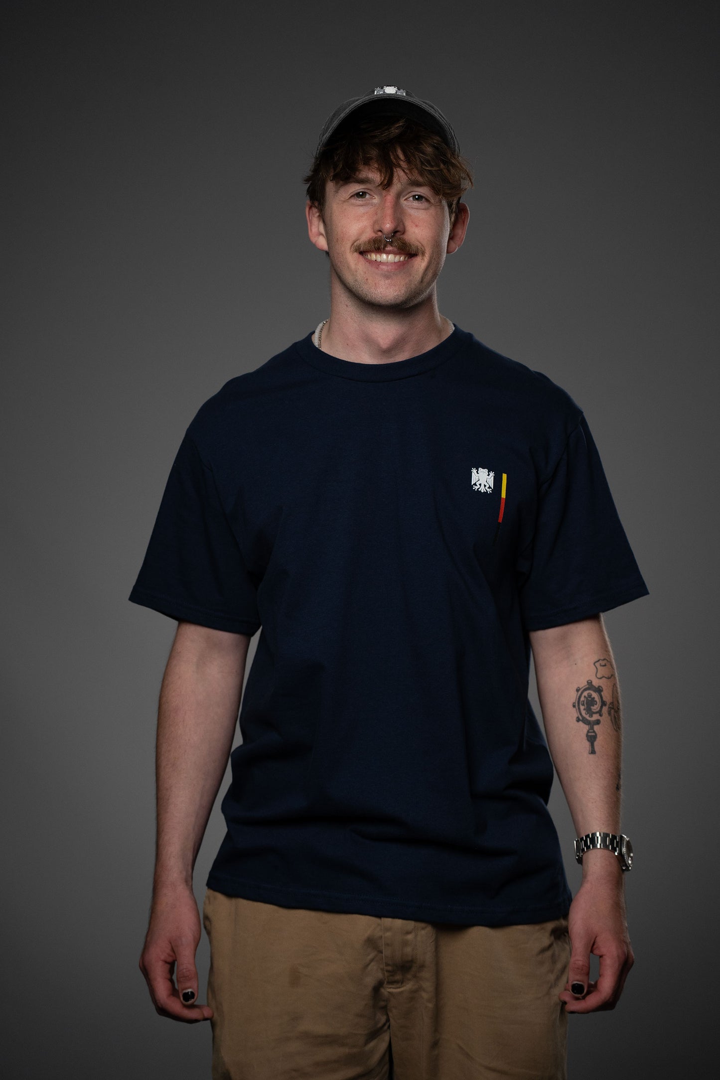 Frontansicht eines dunkelblauen T-Shirts mit kleinem Bundesfrosch-Logo links auf der Brust. Das Model trägt helle Hose und schwarze Kappe, der Fokus liegt auf dem minimalistischen Brustprint.