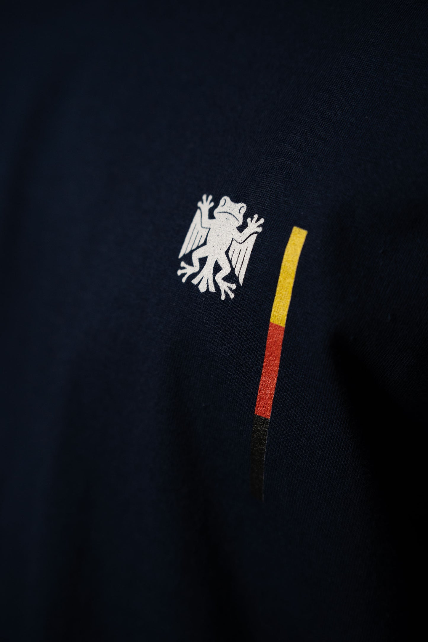 Detailaufnahme des Bundesfrosch-Logos auf dunkelblauem Stoff. Der weiße Frosch mit Flügeln steht neben einem senkrechten Streifen in den Farben Schwarz, Rot und Gelb.