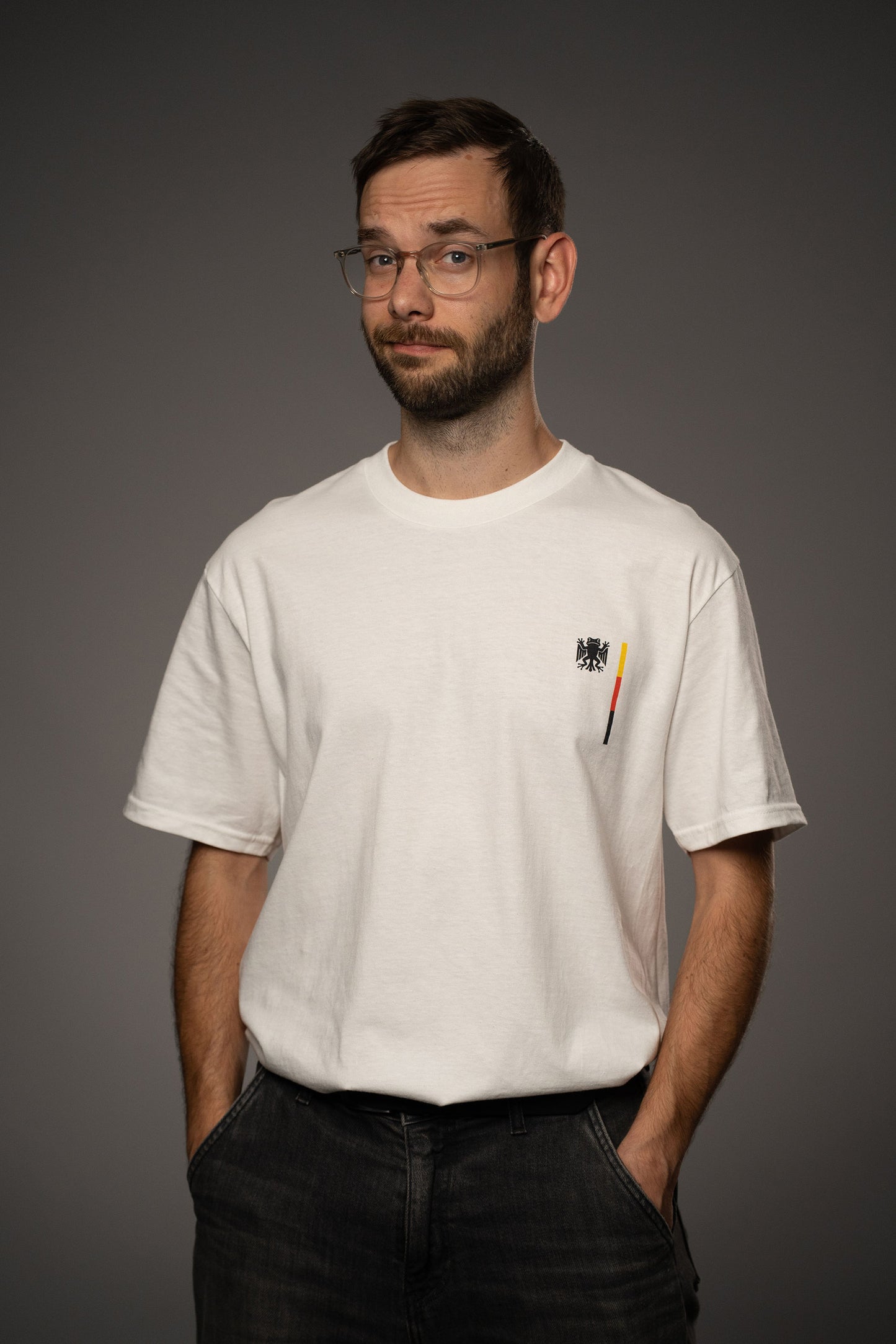 Weißes T-Shirt aus Baumwolle mit kleinem Brustprint „Bundesfrosch“-Logo in Schwarz sowie vertikalem Streifendetail in Schwarz-Rot-Gelb. Das Shirt wird in frontaler Ansicht gezeigt.