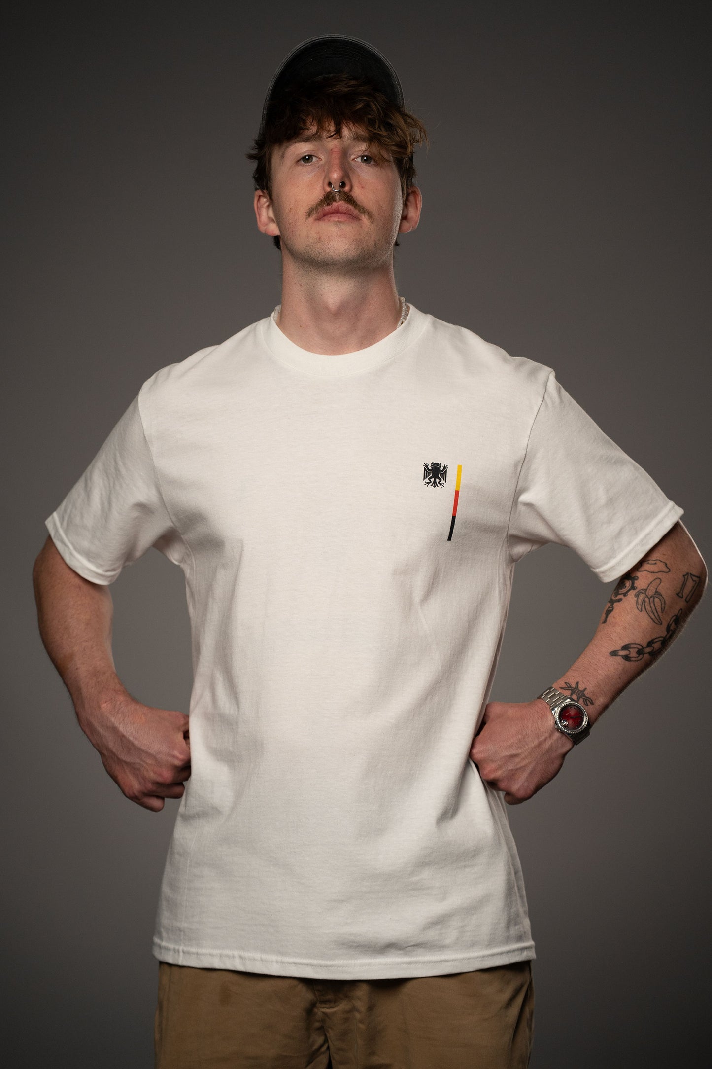 Weißes T-Shirt aus Baumwolle mit schwarzem „Bundesfrosch“-Logo und farbigem Streifenprint in Schwarz-Rot-Gelb auf der Brust. Gezeigt in frontaler Perspektive mit leicht betonter Haltung.