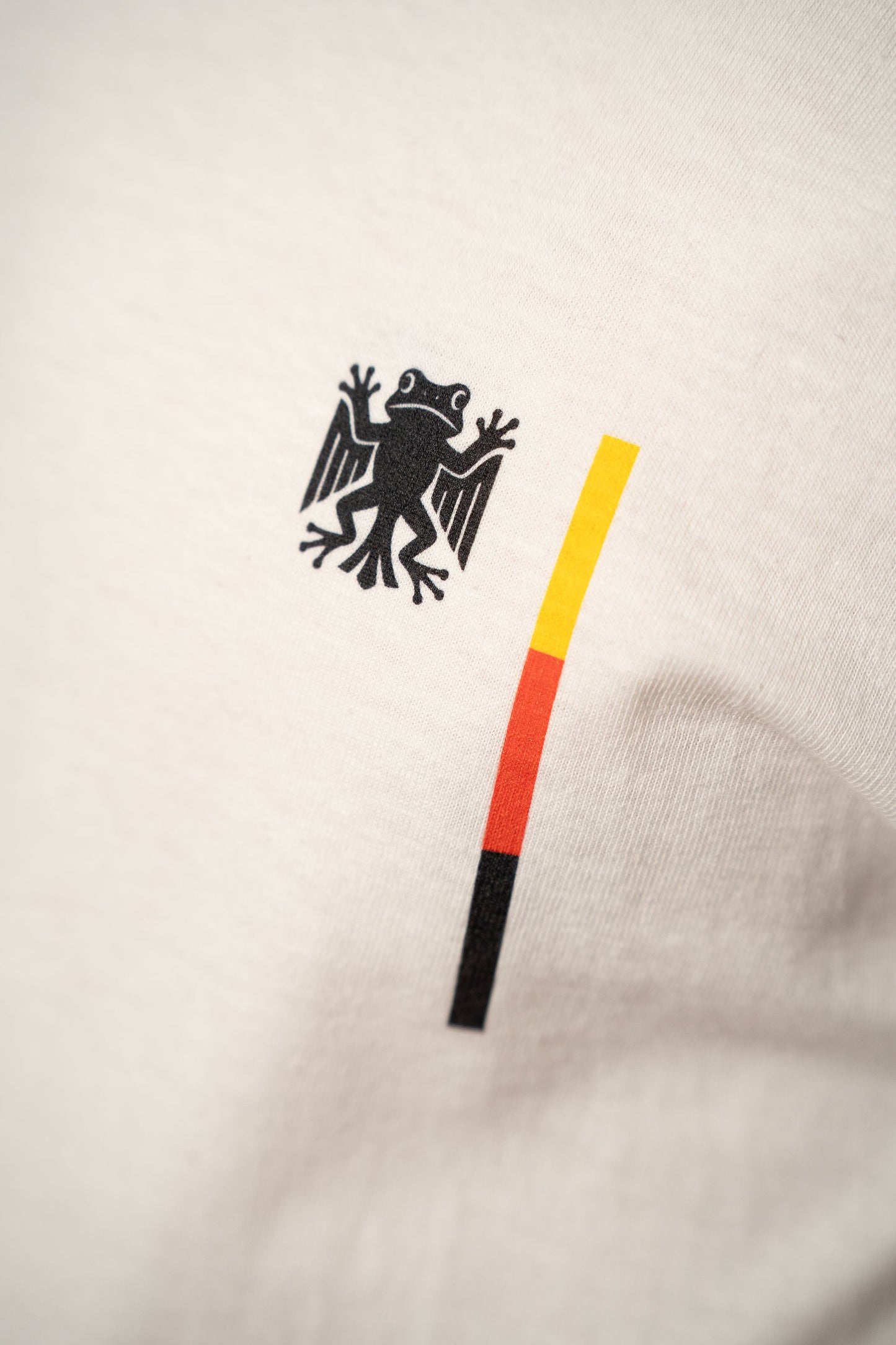 Detailaufnahme des Brustprints auf dem weißen T-Shirt. Zu sehen ist das „Bundesfrosch“-Logo in Schwarz neben einem vertikalen Streifen in den Farben Schwarz, Rot und Gelb.