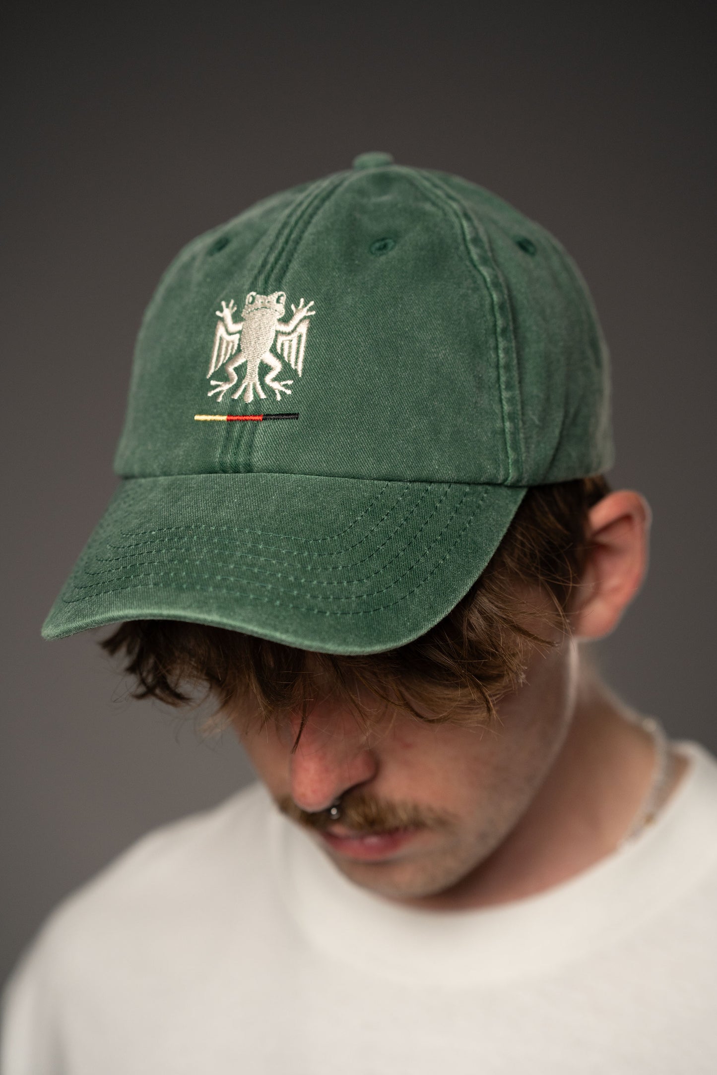 Frontansicht einer grünen Cap aus Baumwollstoff mit weiß gesticktem Bundesfrosch-Logo und kleiner Stickerei in Schwarz, Rot und Gelb unterhalb des Motivs.