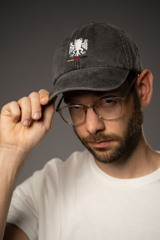 Nahaufnahme einer schwarzen Cap aus Baumwollstoff mit leicht verwaschener Optik. Auf der Vorderseite ist das „Bundesfrosch“-Logo in Weiß gestickt, darunter ein kleiner Streifen in Schwarz-Rot-Gold.