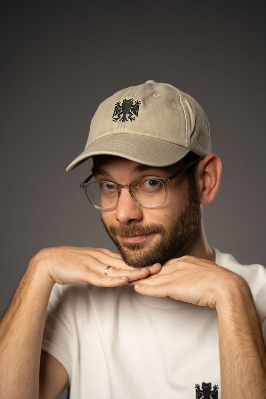 Beigefarbene Baseballcap mit schwarzer Bundesfroschstickerei, getragen von einer Person mit Brille und weißem T-Shirt mit passendem Froschlogo.