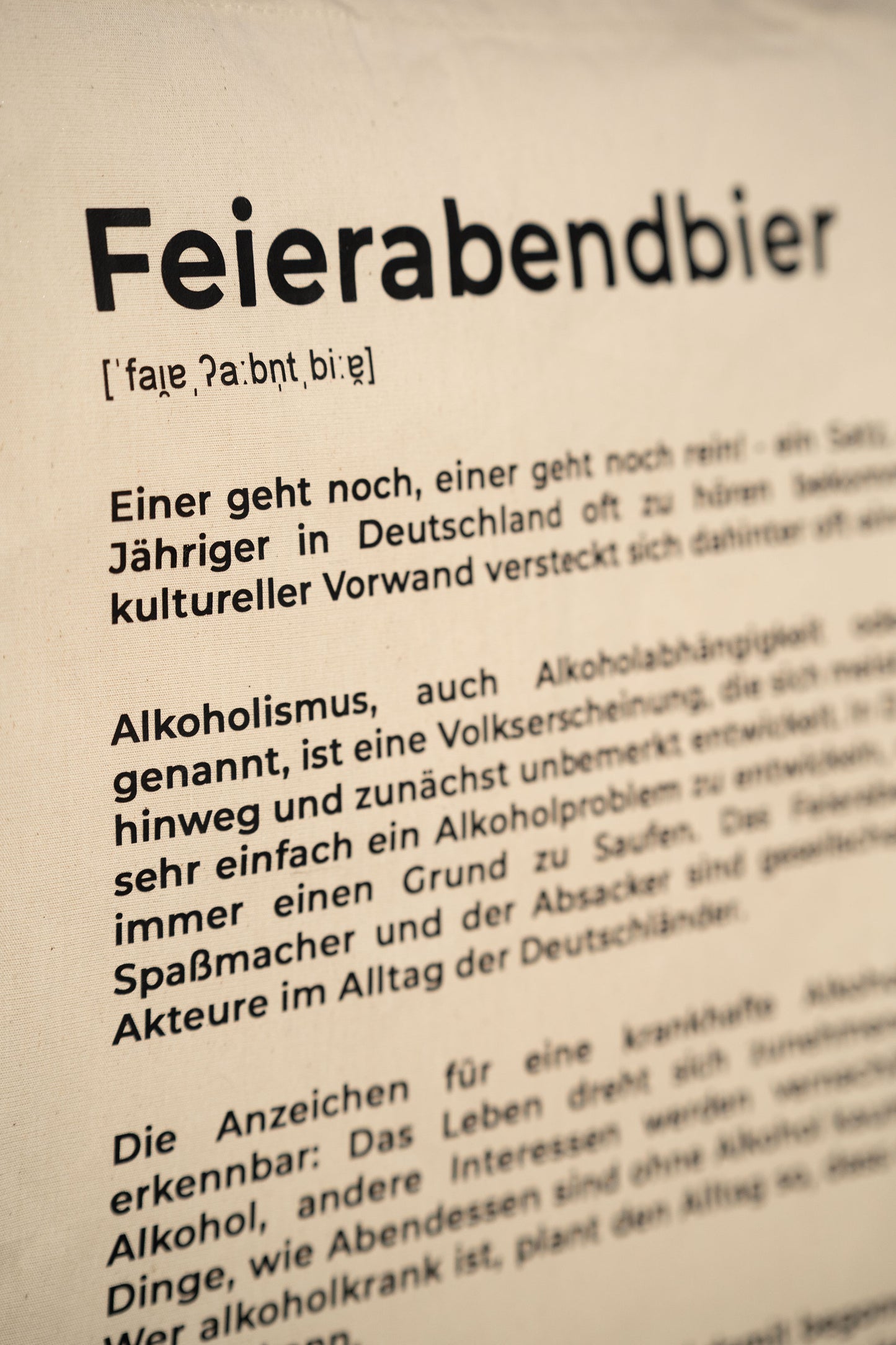 Detailaufnahme der beigefarbenen Stofftasche mit schwarzem Aufdruck „Feierabendbier“ und erklärendem Text im Stil eines Wörterbucheintrags. Der Fokus liegt auf der Typografie und der Textstruktur.