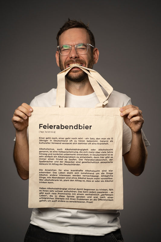 Beigefarbene Stofftasche mit schwarzem Aufdruck „Feierabendbier“ und satirischem Lexikontext über Alkoholkultur in Deutschland. Die Tasche wird von einer Person in weißem T-Shirt hochgehalten, die die Tragehenkel zwischen die Zähne nimmt.