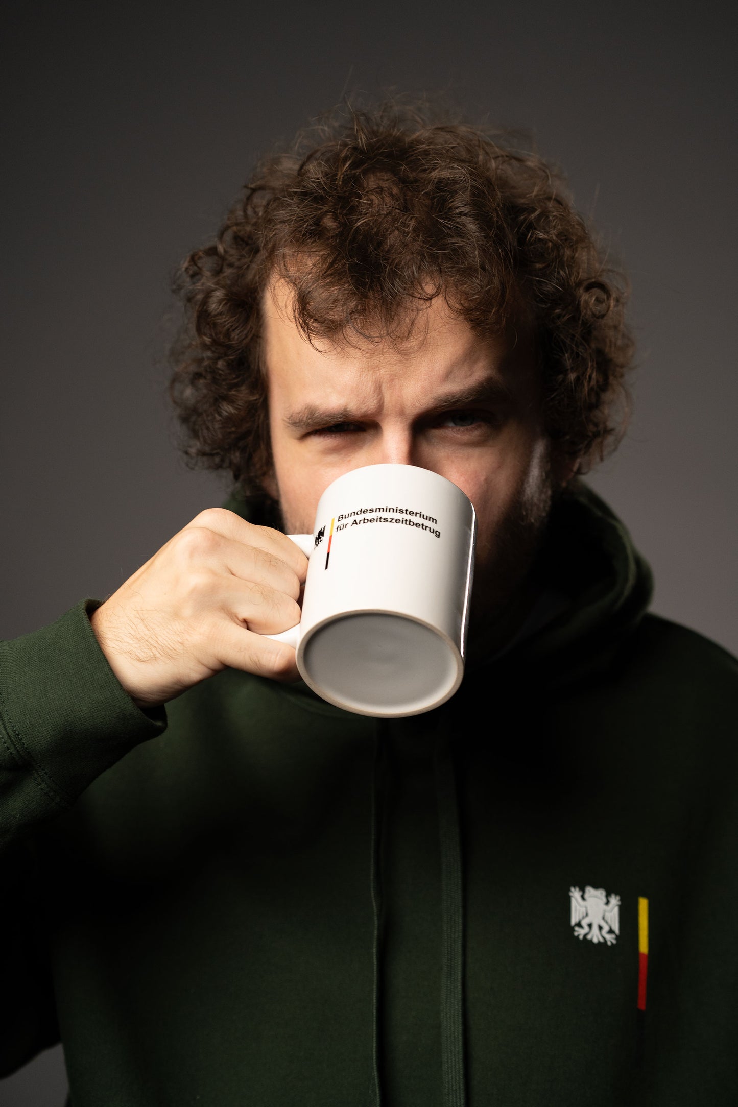 Person in dunkelgrünem Hoodie trinkt aus einer weißen Tasse mit Aufdruck „Bundesministerium für Arbeitszeitbetrug“ und Bundesadler-Logo.