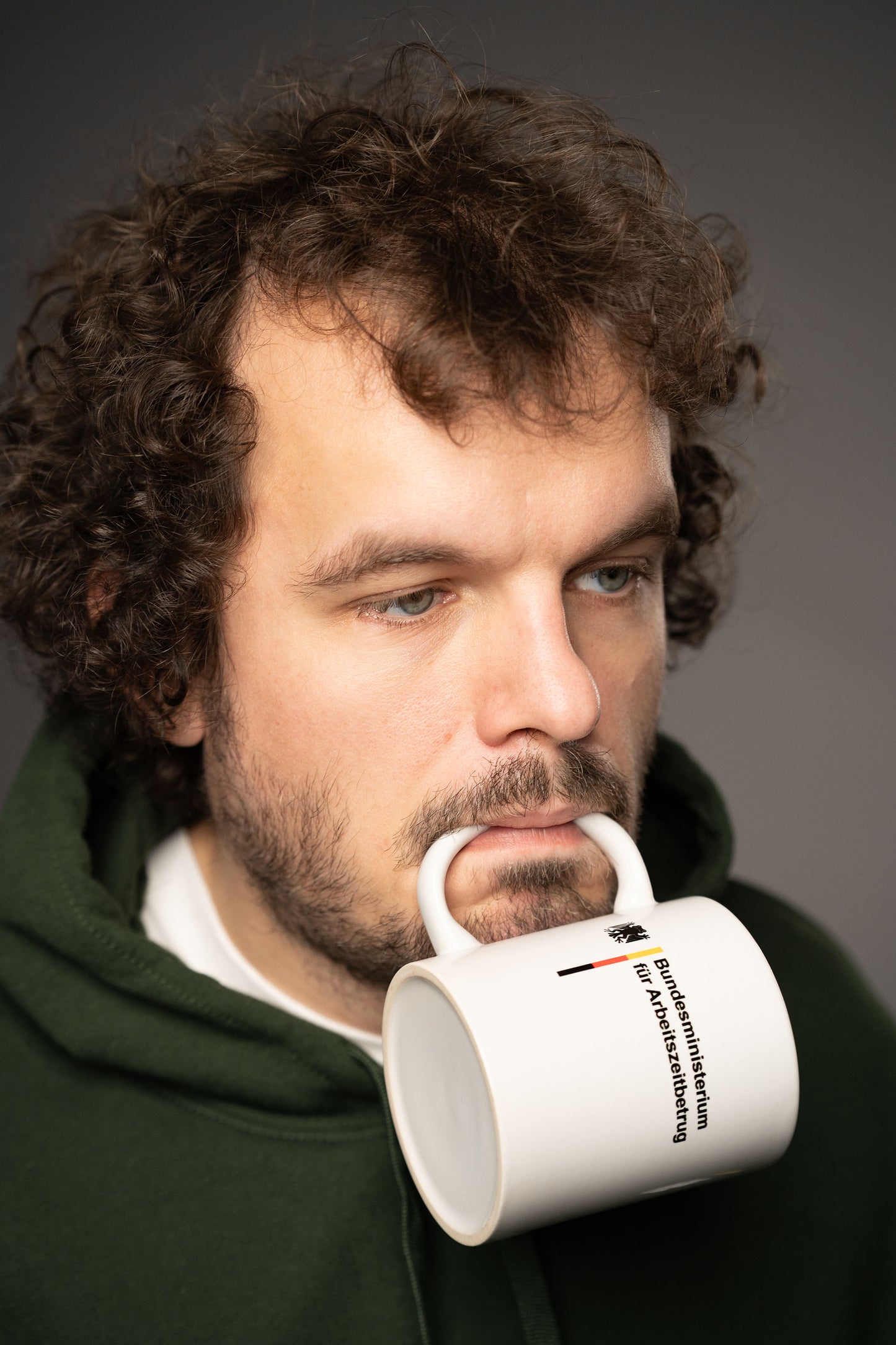 Person mit lockigen Haaren hält eine weiße Tasse mit der Aufschrift „Bundesministerium für Arbeitszeitbetrug“ und Bundesadler-Logo spielerisch im Mund.