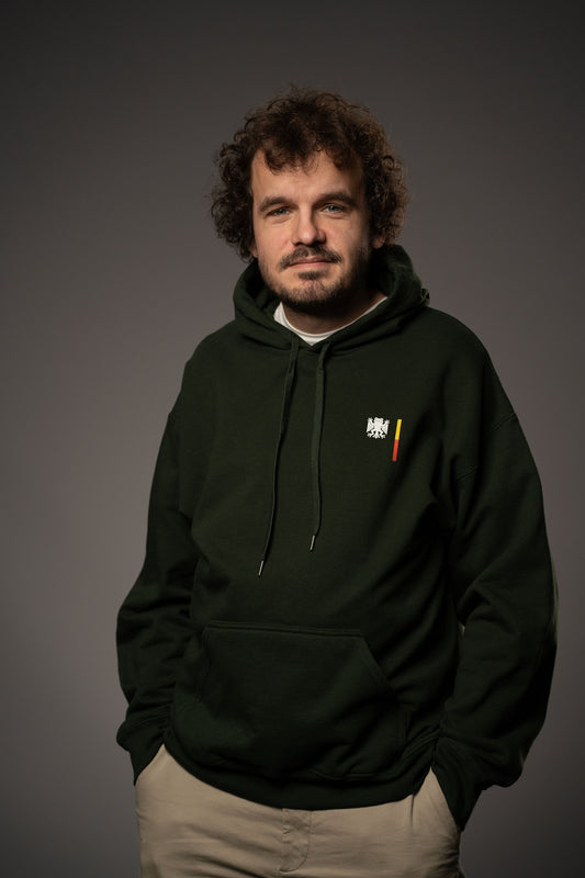 Eine Person trägt einen dunkelgrünen Hoodie mit Brustprint des weißen „Bundesfrosch“-Logos und daneben einem senkrechten Streifen in Schwarz, Rot und Gelb. Der Hoodie hat eine Kängurutasche und Kapuze mit Kordeln.