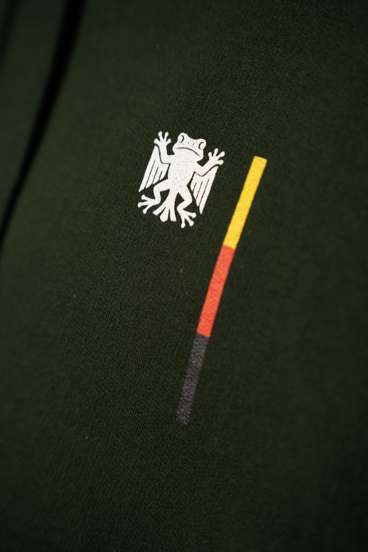Nahaufnahme des Brustprints auf dem dunkelgrünen Hoodie: das weiße „Bundesfrosch“-Logo mit danebenstehendem senkrechtem Balken in den Farben Schwarz, Rot und Gelb.