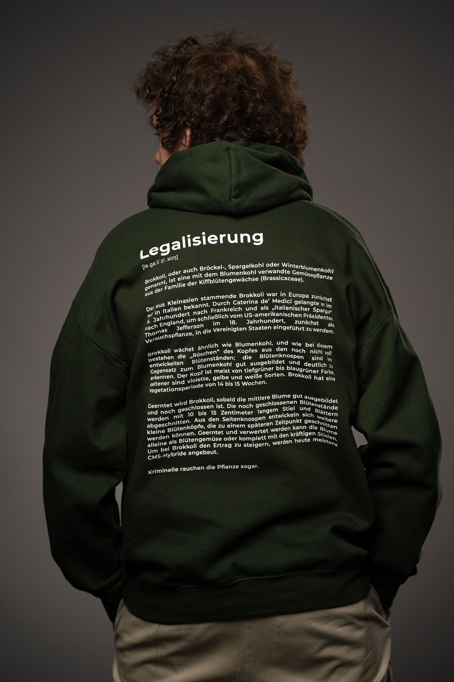 Rückansicht des dunkelgrünen Hoodies mit großem weißen Rückendruck. Oben steht „Legalisierung“ mit phonetischer Schreibweise, darunter ein längerer, textbasierter Druck über Brokkoli in sachlich erklärendem Stil.