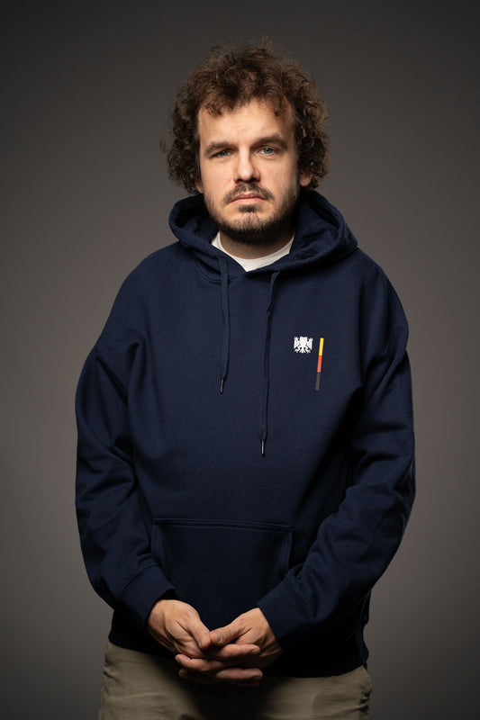 Dunkelblauer Hoodie mit kleiner Brustgrafik in Form des „Bundesfrosch“-Logos – einem stilisierten schwarzen Frosch mit Adlerflügeln – neben einem vertikalen Balken in Schwarz-Rot-Gold. Der Hoodie hat eine Kängurutasche und Kordeln an der Kapuze.