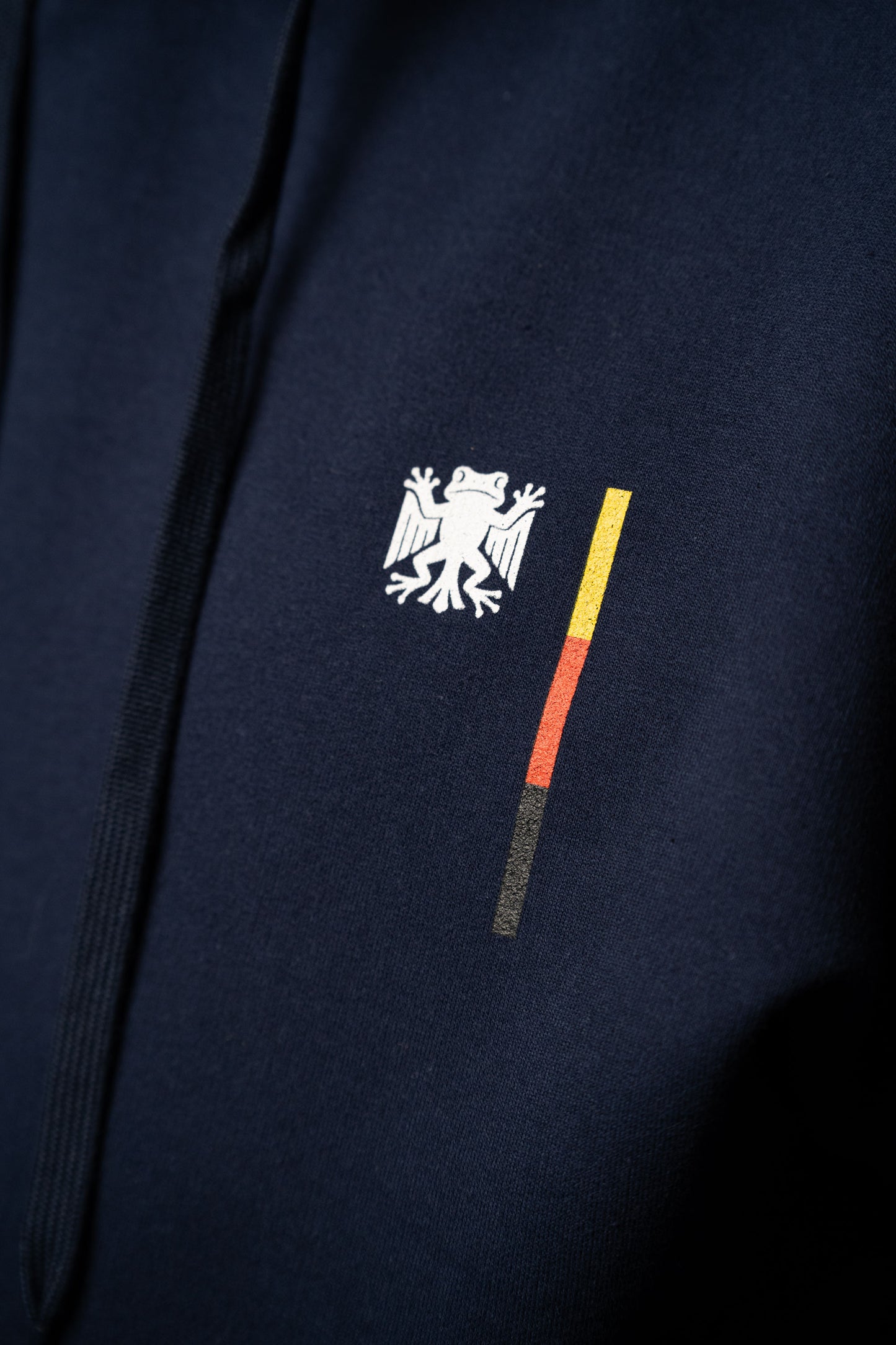 Detailaufnahme der Brustgrafik auf dem dunkelblauen Hoodie: das weiße „Bundesfrosch“-Symbol neben dem vertikalen Balken in Schwarz, Rot und Gelb auf glatter Stoffoberfläche.