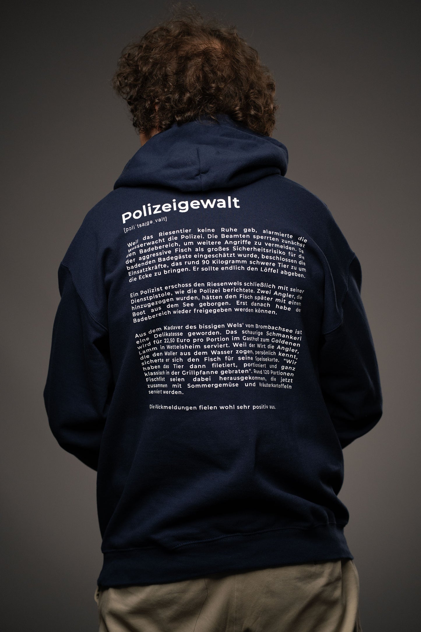 Rückansicht des dunkelblauen Hoodies mit großem weißen Textprint. Der Aufdruck trägt die Überschrift „Polizeigewalt“ mit Lautschrift und einem satirischen Fließtext im Lexikonstil.