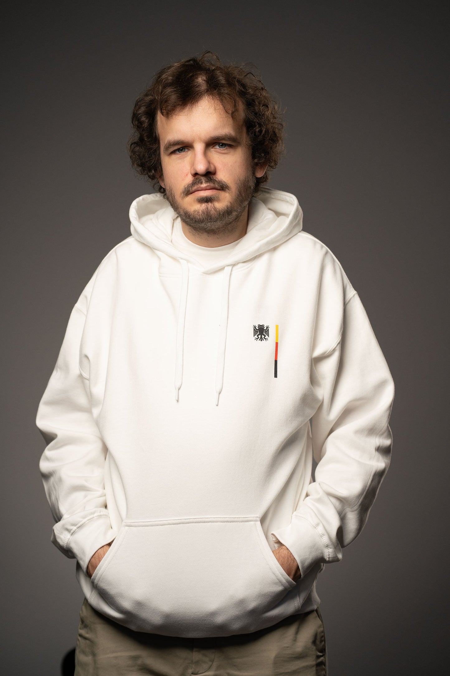 Person trägt einen weißen Hoodie mit kleinem Brustprint. Auf der linken Brust ist das Logo „Bundesfrosch“ in Schwarz mit einem vertikalen Streifen in Schwarz, Rot und Gelb aufgedruckt.
