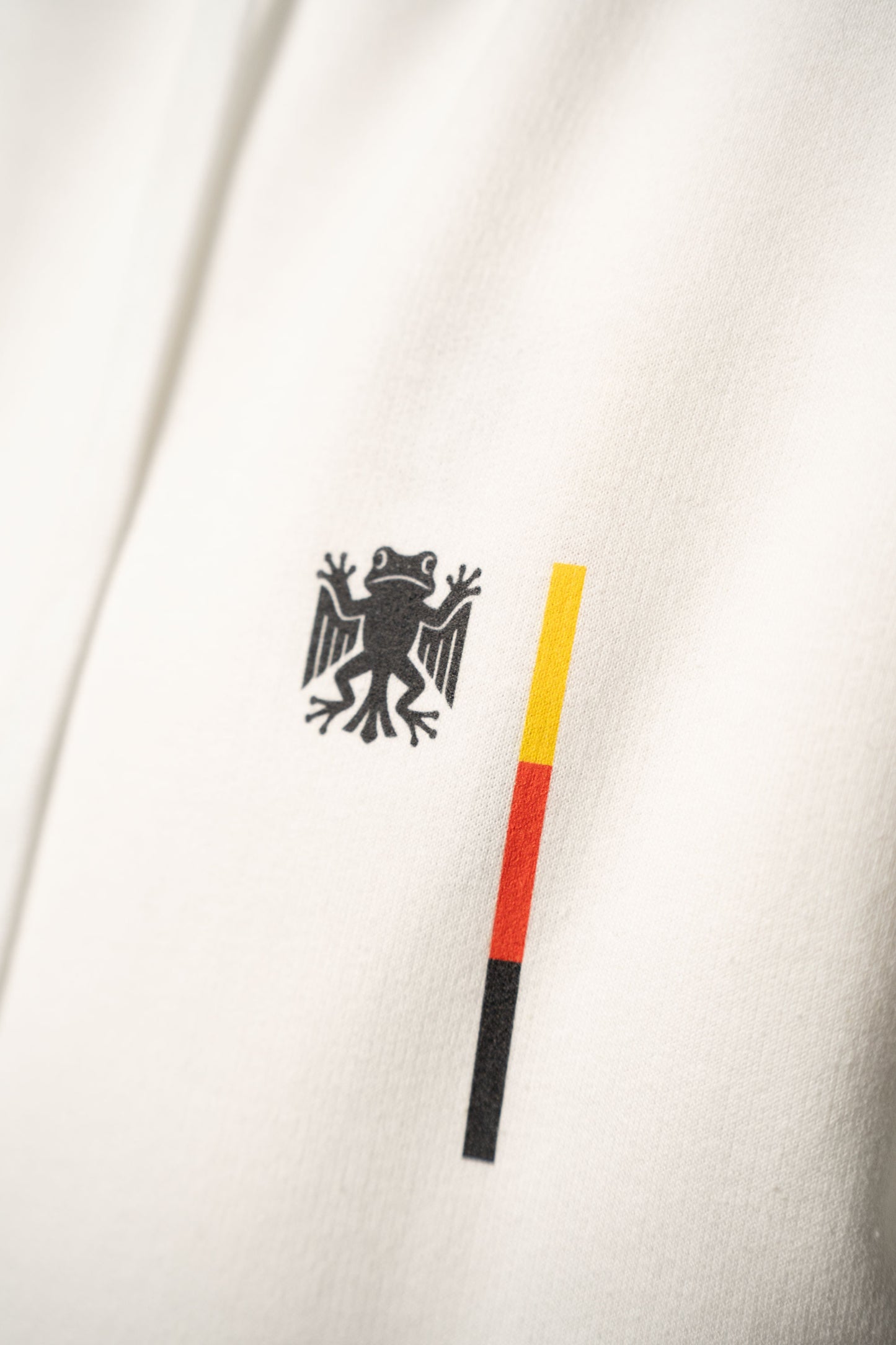 Nahaufnahme des Brustprints auf dem weißen Hoodie. Sichtbar ist das schwarze „Bundesfrosch“-Logo neben einem schmalen Balken in den Farben Schwarz, Rot und Gelb.