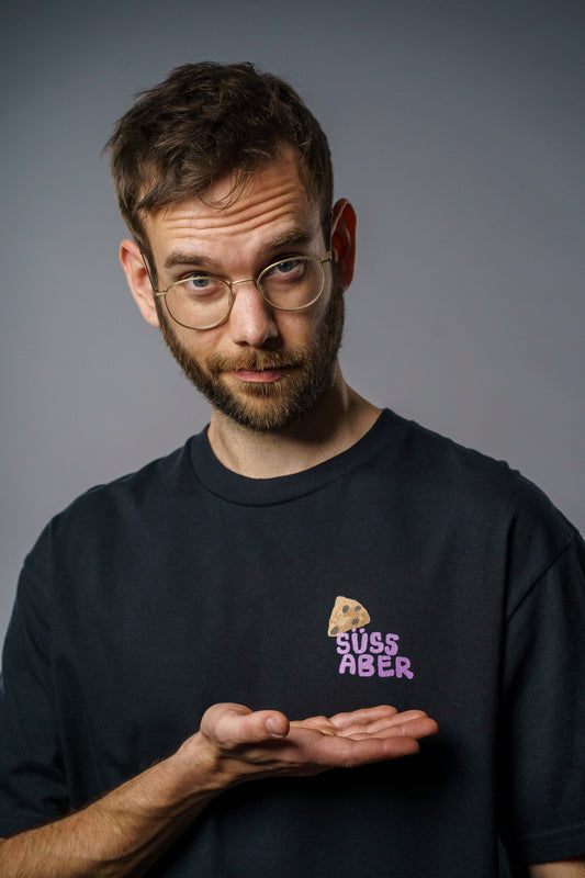 Schwarzes T-Shirt mit kleinem Brust-Print: Ein gezeichneter Cookie über dem Schriftzug „SÜSS ABER“ in kräftigem Lila. Das Design sitzt auf der linken Brustseite und kombiniert humorvolle Typografie mit schlichtem Stil. Das Shirt hat einen klassischen Rundhalsausschnitt und eine lockere Passform.