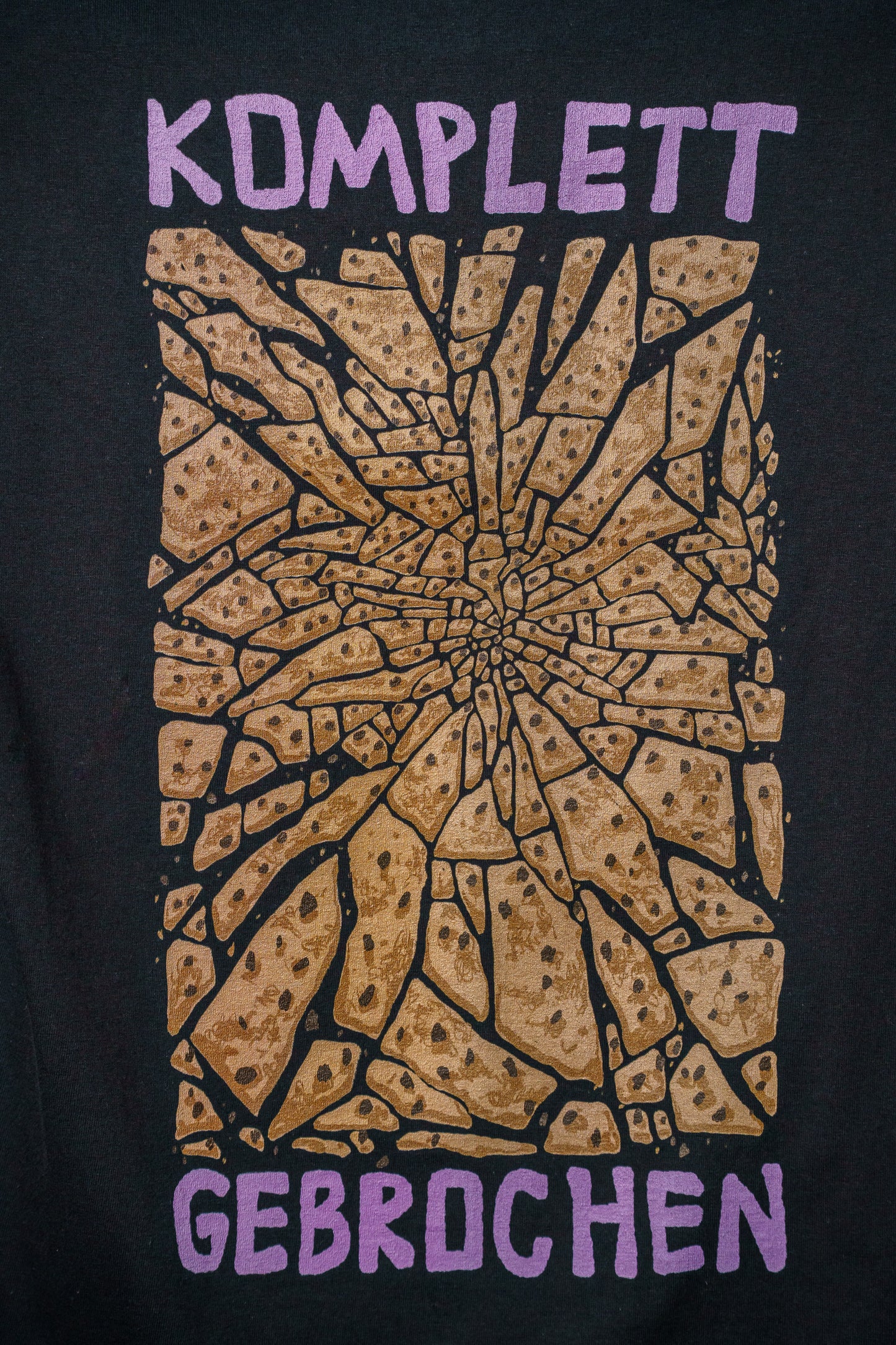 Großer Rückenprint auf einem schwarzen T-Shirt: In der Mitte ein bräunlich-gelbes, splitterartiges Muster mit dunklen Punkten – stilisiert wie zerbrochene Keksstücke. Darüber in kräftigem Lila der Schriftzug „KOMPLETT“, darunter „GEBROCHEN“, jeweils in handschriftlich wirkender Blockschrift. Das Motiv kombiniert Humor mit starker grafischer Wirkung.