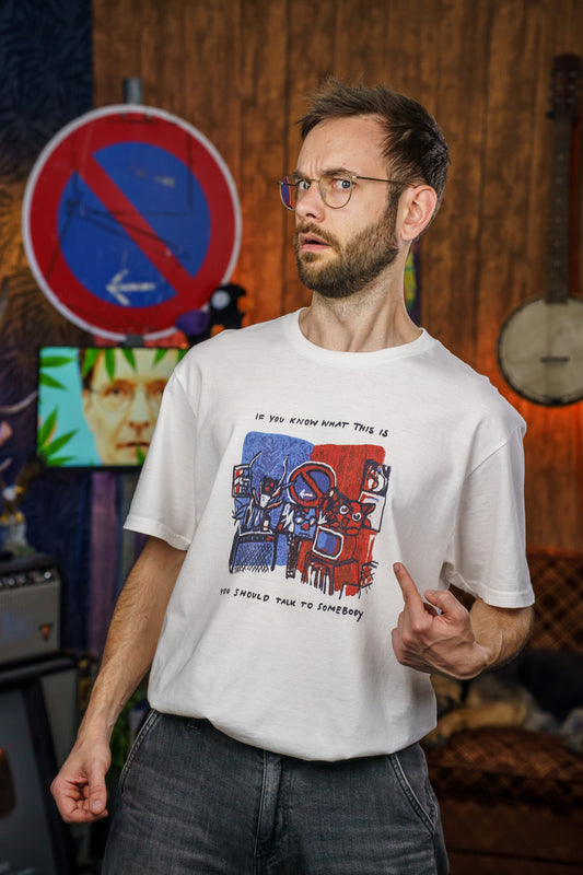 Ein Mann mit Brille und Bart steht in einem Raum und zeigt mit dem Finger auf sein weißes T-Shirt. Das Shirt zeigt eine bunte, abstrakte Illustration in Rot und Blau mit mehreren stilisierten Figuren und Fernsehern. Darüber steht der Text „If you know what this is“, darunter „you should talk to somebody“. Der Mann blickt mit fragender oder skeptischer Miene zur Seite. Im Hintergrund sind unter anderem ein Verbotsschild, ein Banjo und ein Bildschirm mit einem Gesicht zu sehen.