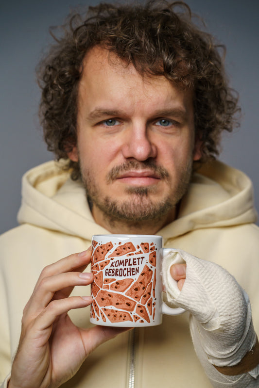 Ein Mann in cremefarbenem Hoodie hält eine weiße Keramiktasse mit beiden Händen vor sich. Die Tasse trägt ein braun-orangenes, rissiges Muster und in der Mitte den Schriftzug „KOMPLETT GEBROCHEN“. Seine rechte Hand ist dick bandagiert. Der Mann blickt neutral bis ernst in die Kamera.