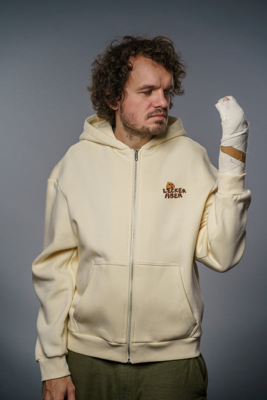 Ein Mann mit lockigen Haaren trägt einen cremefarbenen Zip-Hoodie mit der kleinen Aufschrift „Lecker aber“ und einem Keks-Motiv auf der linken Brustseite. Er blickt ernst auf seine rechte Hand, die dick mit Verband umwickelt ist. Der Hintergrund ist neutral grau.
