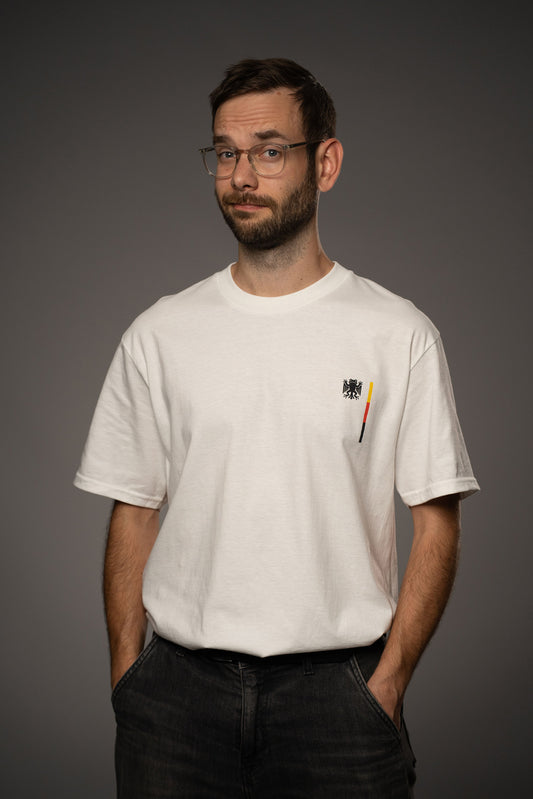 Weißes T-Shirt aus Baumwolle mit kleinem Brustprint „Bundesfrosch“-Logo in Schwarz sowie vertikalem Streifendetail in Schwarz-Rot-Gelb. Das Shirt wird in frontaler Ansicht gezeigt.