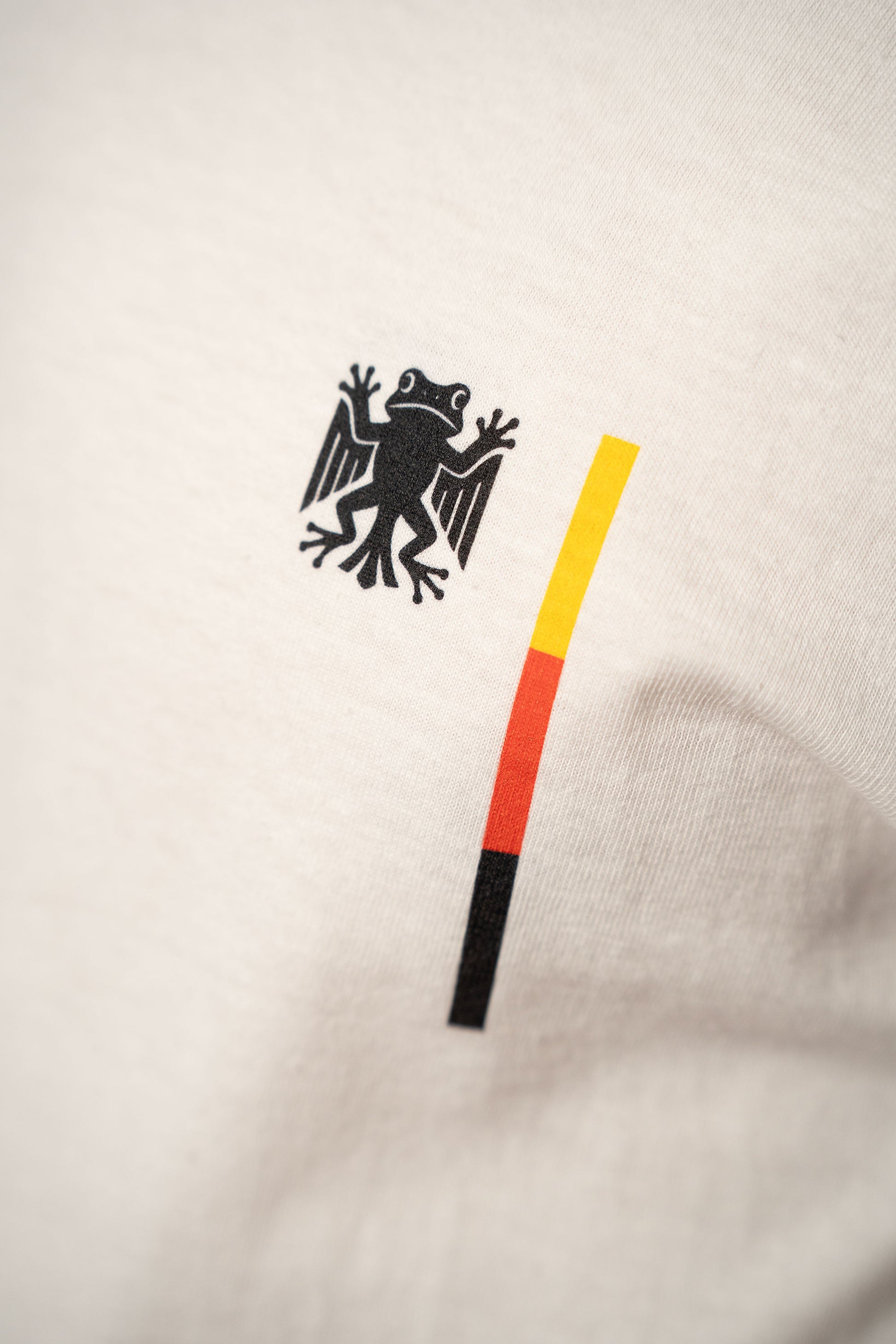 Detailaufnahme des Brustprints auf dem weißen T-Shirt. Zu sehen ist das „Bundesfrosch“-Logo in Schwarz neben einem vertikalen Streifen in den Farben Schwarz, Rot und Gelb.