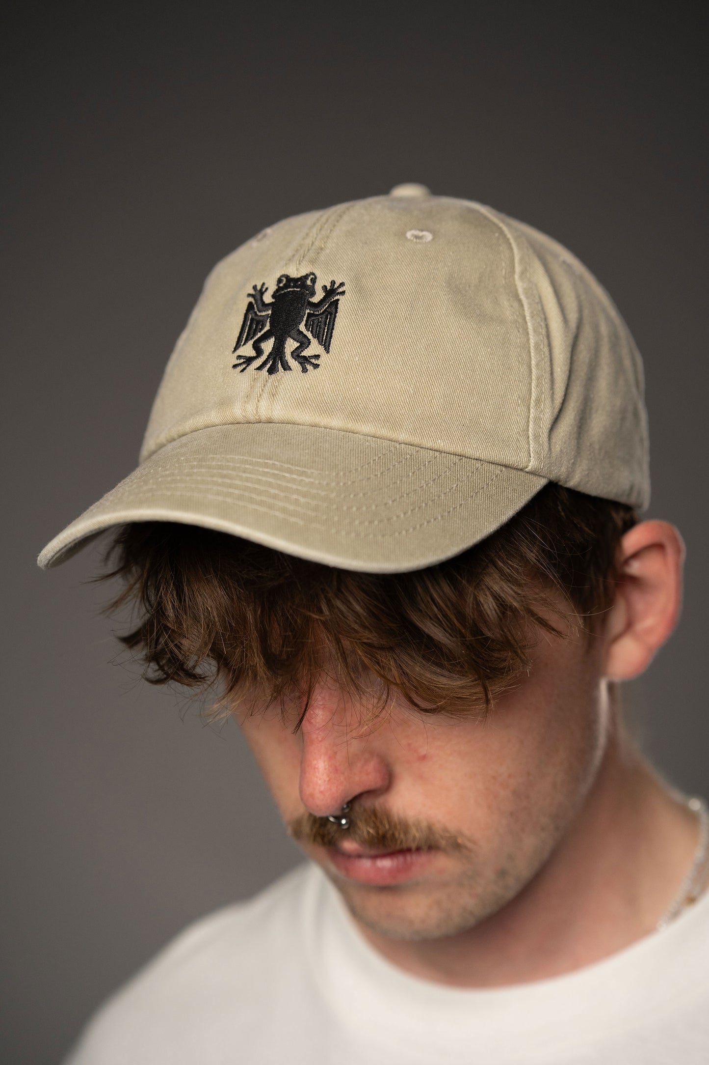 Beigefarbene Baseballcap mit schwarzer Stickerei eines stilisierten Frosches auf der Vorderseite, getragen von einer Person in weißem T-Shirt.