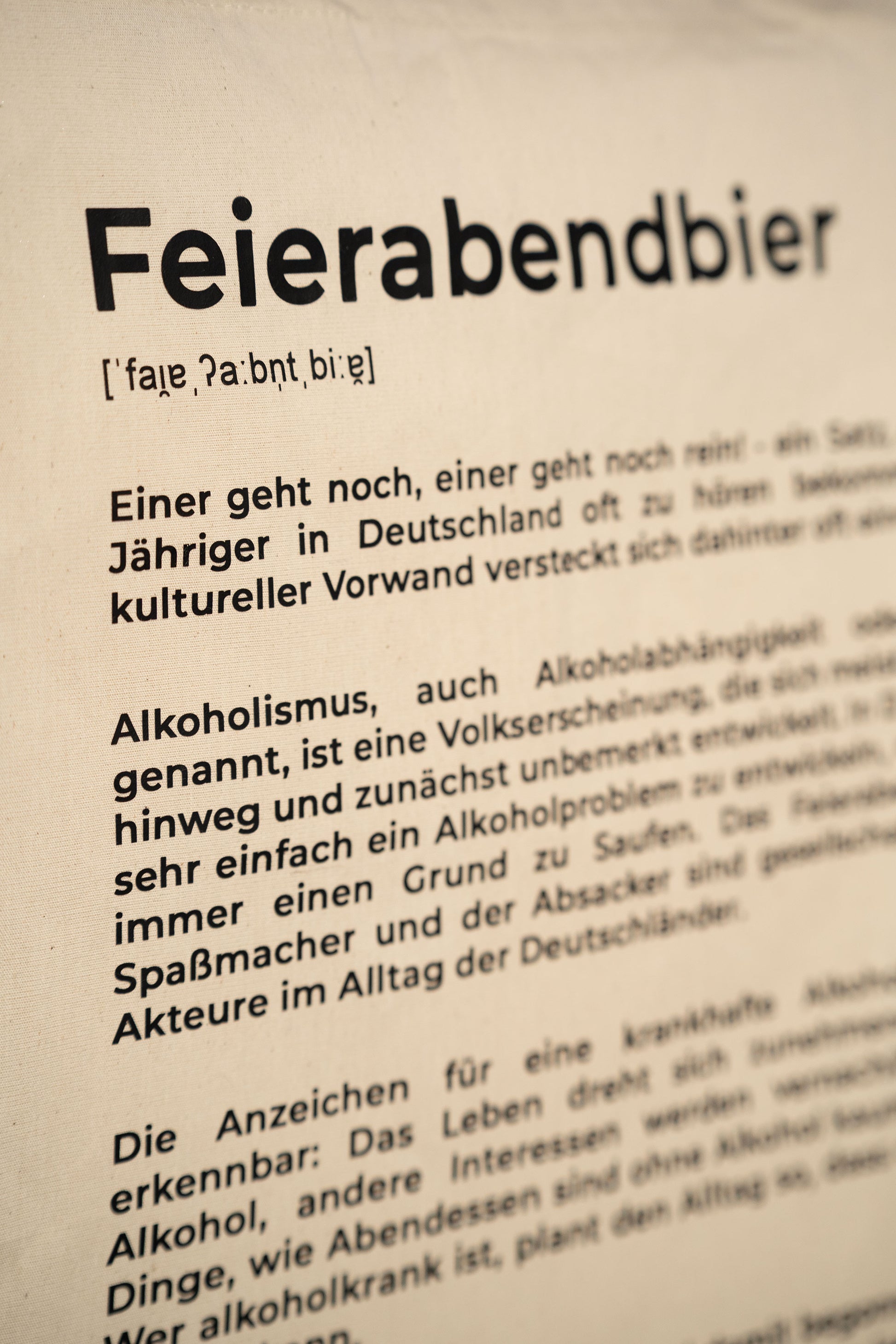 Detailaufnahme der beigefarbenen Stofftasche mit schwarzem Aufdruck „Feierabendbier“ und erklärendem Text im Stil eines Wörterbucheintrags. Der Fokus liegt auf der Typografie und der Textstruktur.