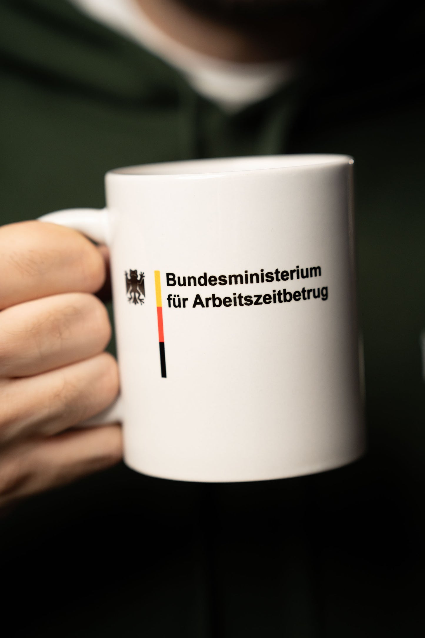 Weiße Keramiktasse mit Aufdruck „Bundesministerium für Arbeitszeitbetrug“ und kleinem Bundesadler-Logo, gehalten in einer Hand.