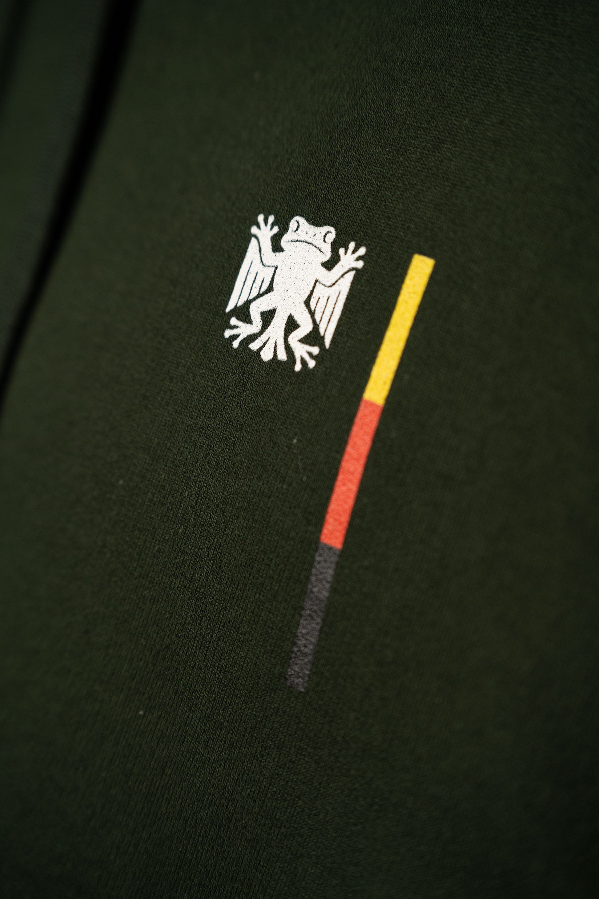 Nahaufnahme des Brustprints auf dem dunkelgrünen Hoodie: das weiße „Bundesfrosch“-Logo mit danebenstehendem senkrechtem Balken in den Farben Schwarz, Rot und Gelb.