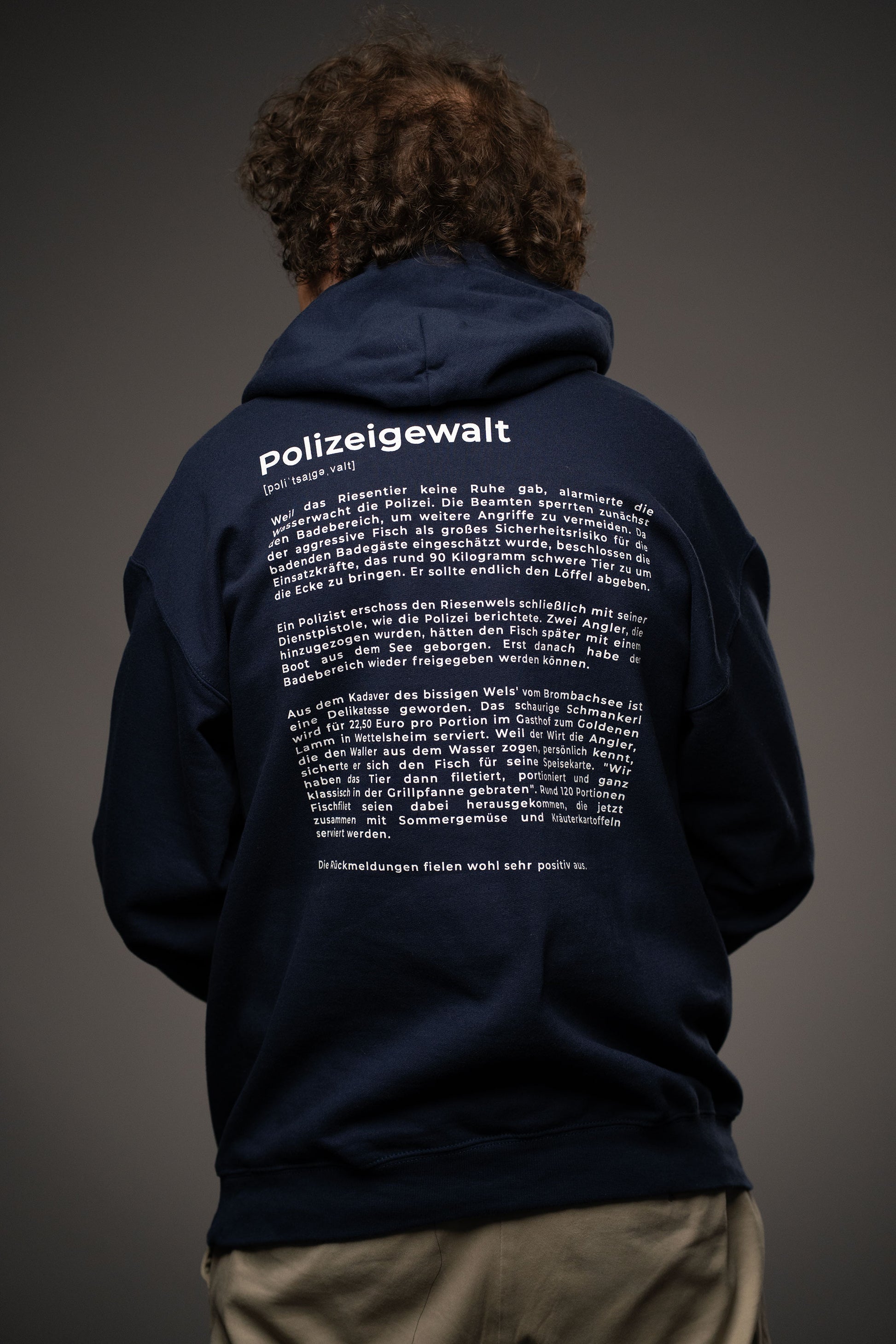 Rückansicht des dunkelblauen Hoodies mit großem weißen Textprint. Der Aufdruck trägt die Überschrift „Polizeigewalt“ mit Lautschrift und einem satirischen Fließtext im Lexikonstil.