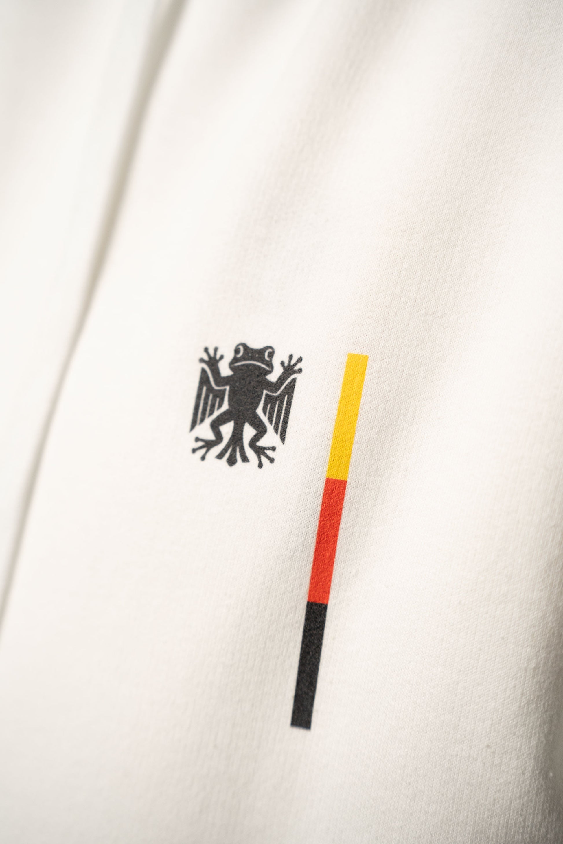 Nahaufnahme des Brustprints auf dem weißen Hoodie. Sichtbar ist das schwarze „Bundesfrosch“-Logo neben einem schmalen Balken in den Farben Schwarz, Rot und Gelb.
